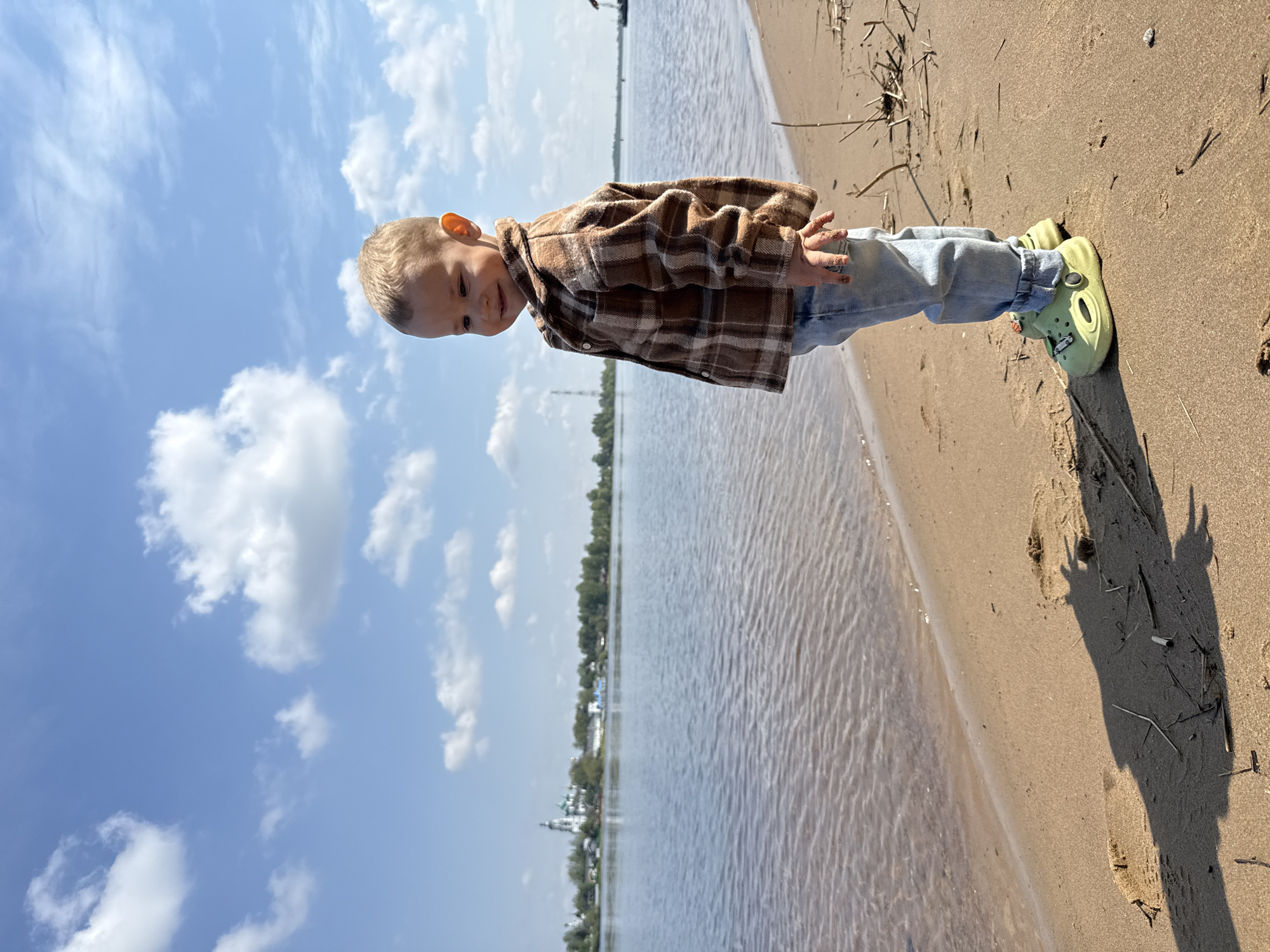 A toddler stands on the sandy shore of a body of water under a blue, cloudy sky. The child wears a brown and white plaid shirt, blue jeans, and green Crocs. The sand is a light beige color. In the background, across the water, is a line of trees and buildings. The image was taken in Rusanovo, Russia.