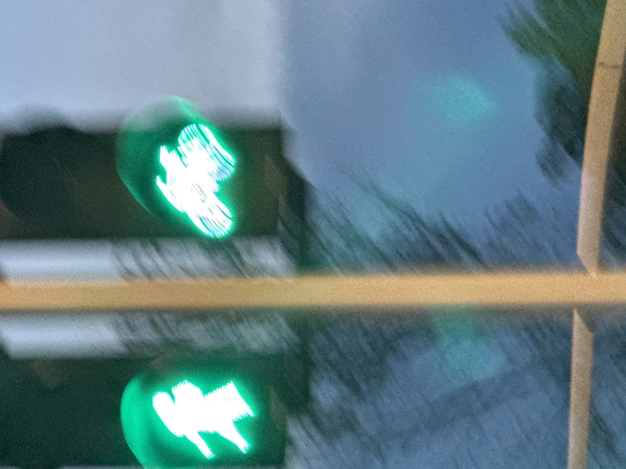 The image shows a blurred view of two green traffic signals. The signal on the left displays a white silhouette of a pedestrian in a walking pose, indicating it is safe to cross. The signal on the right shows a white silhouette of a motorcycle, suggesting a green light for vehicular traffic. The signals are illuminated against a dark background, with a vertical yellow pole in between them. The overall scene is indistinct due to motion blur, suggesting the photograph was taken while in motion. The environment appears to be outdoors in an urban setting, with diffused lighting that suggests it is either dusk or an overcast day. There are no discernible people or other specific objects in the background, only abstract blurs of color and light.