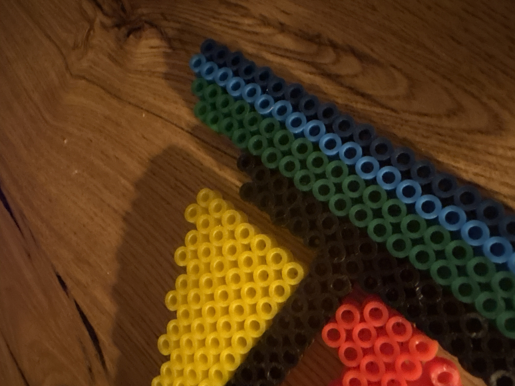 The image shows a close-up, overhead view of several rows of colorful plastic beads arranged on a wooden surface. The beads are small, circular, and hollow, resembling perler beads. They are arranged in distinct color blocks and diagonal lines. The colors visible are yellow, black, red, green, and blue. The yellow beads form a triangular shape. The red beads are clustered in a partial shape resembling a heart or a cluster of flowers. The black beads are in a solid row. The green and blue beads are arranged in parallel diagonal rows, creating a striped effect. The wooden surface has a visible grain pattern and appears to be a table or floor. Shadows are cast on the wooden surface by the beads, indicating a light source from the upper left. There are no people or animals present in the image. The image does not provide any information about the location of Ореховка, Russia.