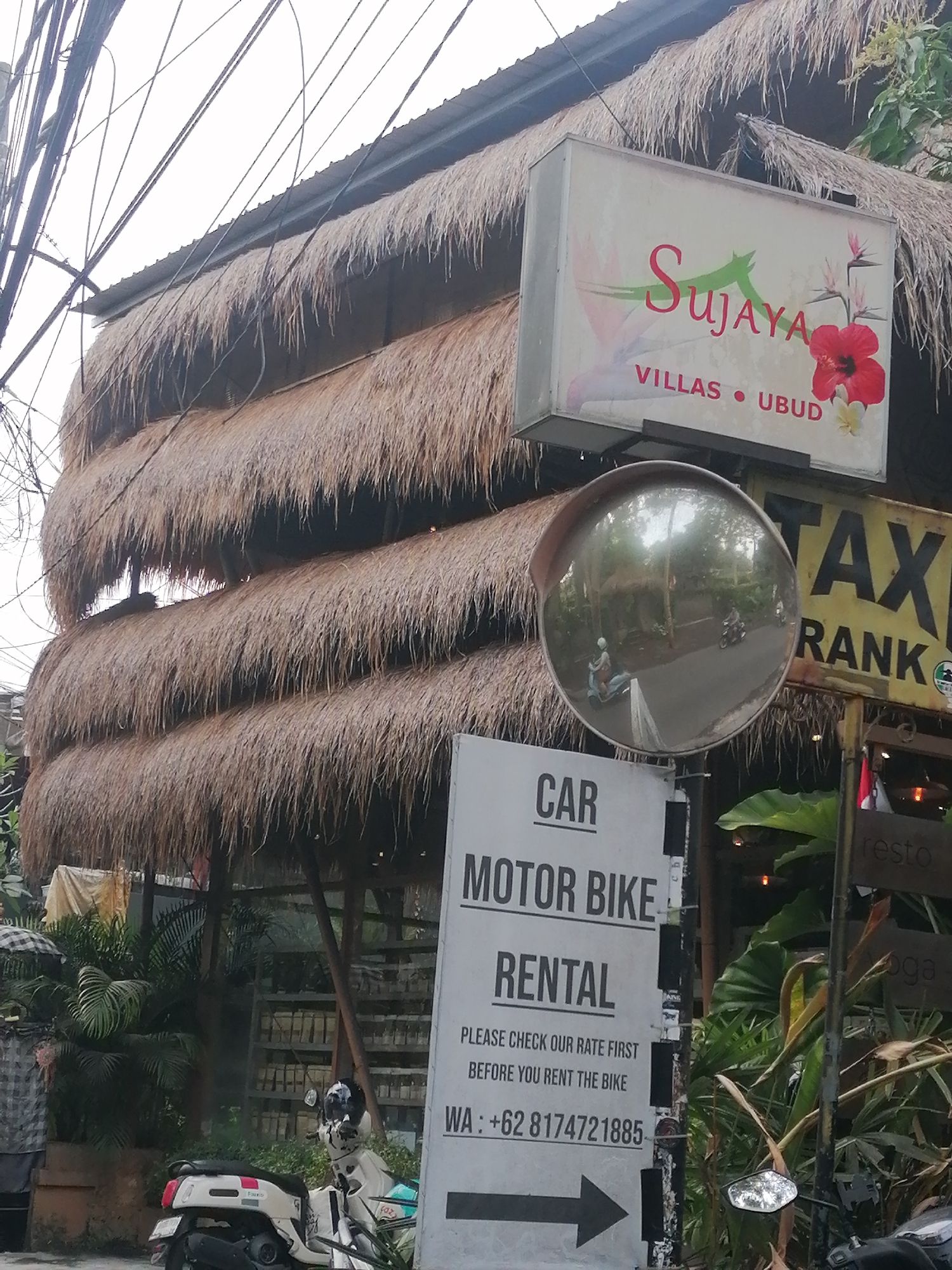 This image depicts a street scene in the Ubud District, Indonesia, featuring a building with a thatched roof and several signs. The building appears to be a rental establishment, possibly for villas or motorbikes. A prominent sign reads "SUJAYA VILLAS - UBUD" with hibiscus flowers. Another sign indicates "CAR MOTOR BIKE RENTAL," with contact information provided as a WhatsApp number. In the background, a yellow "TAXI RANK" sign is visible. A convex mirror is mounted on a pole, reflecting the street with passing motorbikes. Lush green foliage and palm trees are present, characteristic of the tropical environment. A white scooter is parked in the foreground. Overhead, a complex network of electrical wires crisscrosses the sky.