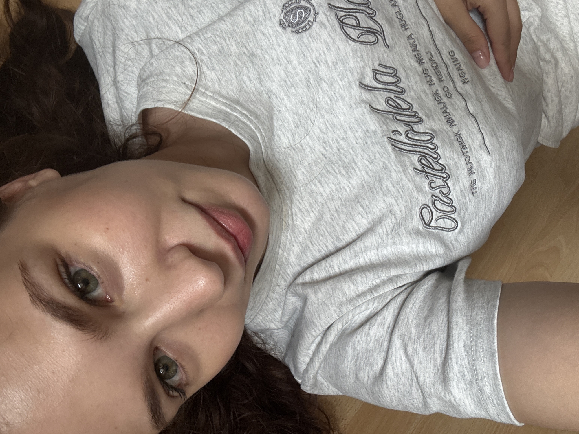 The image shows a close-up, overhead shot of a young woman lying on a wooden floor. She is wearing a light grey t-shirt with embroidered text and a logo on it. Her face is tilted upwards, with her eyes, nose, and mouth in focus. Her eyes are green, and her lips are slightly parted, appearing to be wearing lip gloss. Her dark brown hair is spread around her head and shoulders. A hand with manicured nails is visible in the upper left corner of the image, resting on the t-shirt. The background is a plain wooden surface, suggesting an indoor setting. There is no discernible indication of the city of Chaikovsky, Russia, within the image.