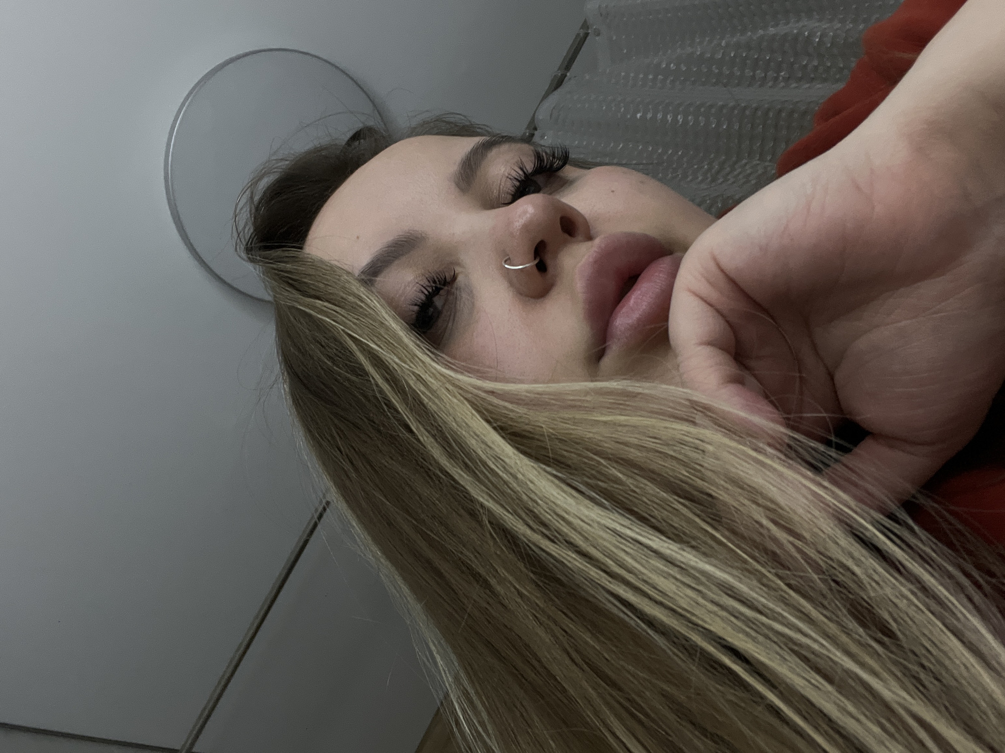 The image shows a woman with long, light brown hair. She has a silver nose ring and visible eyelashes. Her face rests on her hand. The background is blurry but appears to be a light-colored wall with a round mirror. The location is not discernible from the image; there is no indication that this image was taken in the city of Krasnodar, Russia.