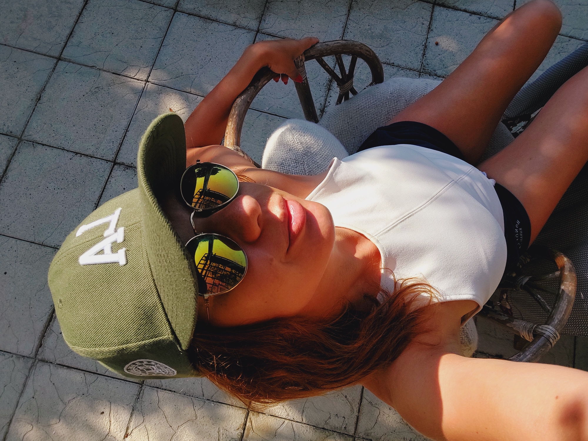 A woman sits outdoors on a tiled surface. She is wearing a green baseball cap with white lettering, sunglasses with mirrored lenses, and a white sleeveless top over dark shorts. Her right arm rests on the back of a chair. The scene appears to be illuminated by sunlight, casting shadows. The location is the city of Sozopol, Bulgaria.