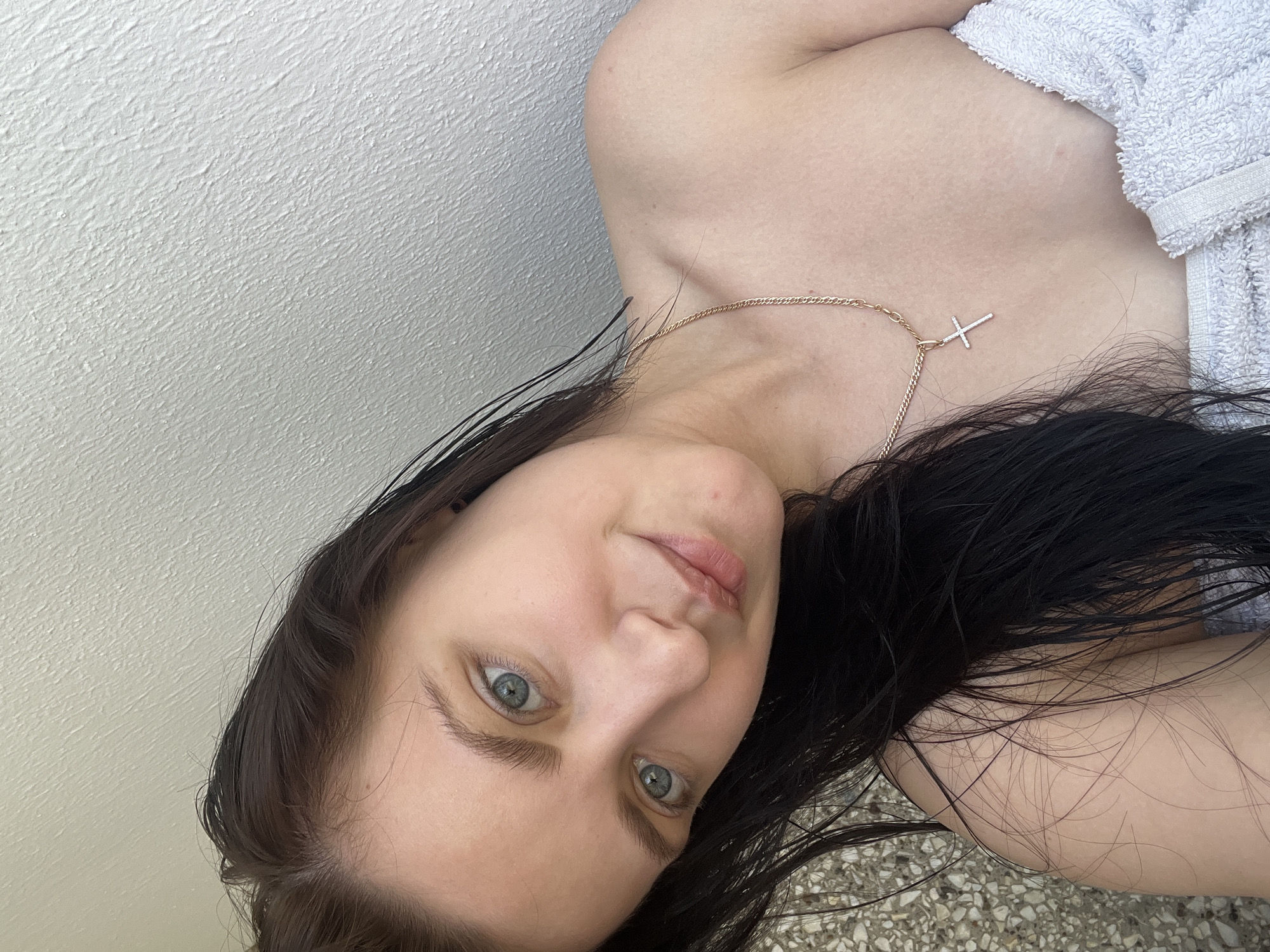 A woman lies on her side, partially covered by a white towel. She is wearing a gold necklace with a cross pendant. Her hair is dark and appears wet. She has fair skin and light blue eyes. The background suggests she is near a white wall and some kind of tiled or textured surface. The location given is Марковский, Russia.