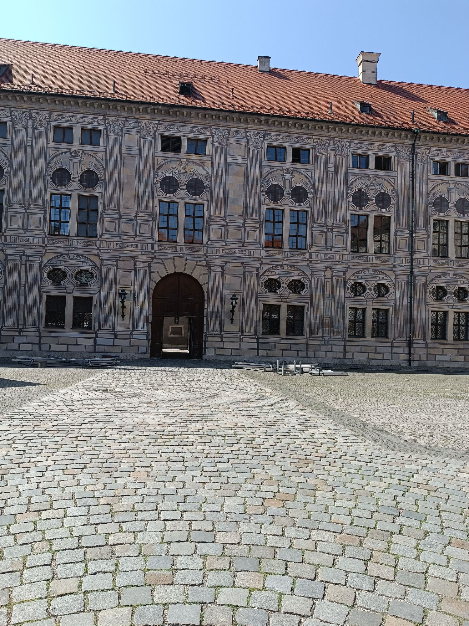 The image depicts the courtyard of the Residenz in Munich, Germany. The Residenz is a former royal palace of the Bavarian monarchs. The photograph shows a cobblestone courtyard in the foreground, leading towards the grand facade of the palace. The palace building is a large, ornate structure with a red tiled roof. It features numerous windows, decorative architectural details, and a large arched wooden doorway in the center. The sky is clear and blue, indicating a sunny day. There are no people visible in the image, and the scene is calm and still.