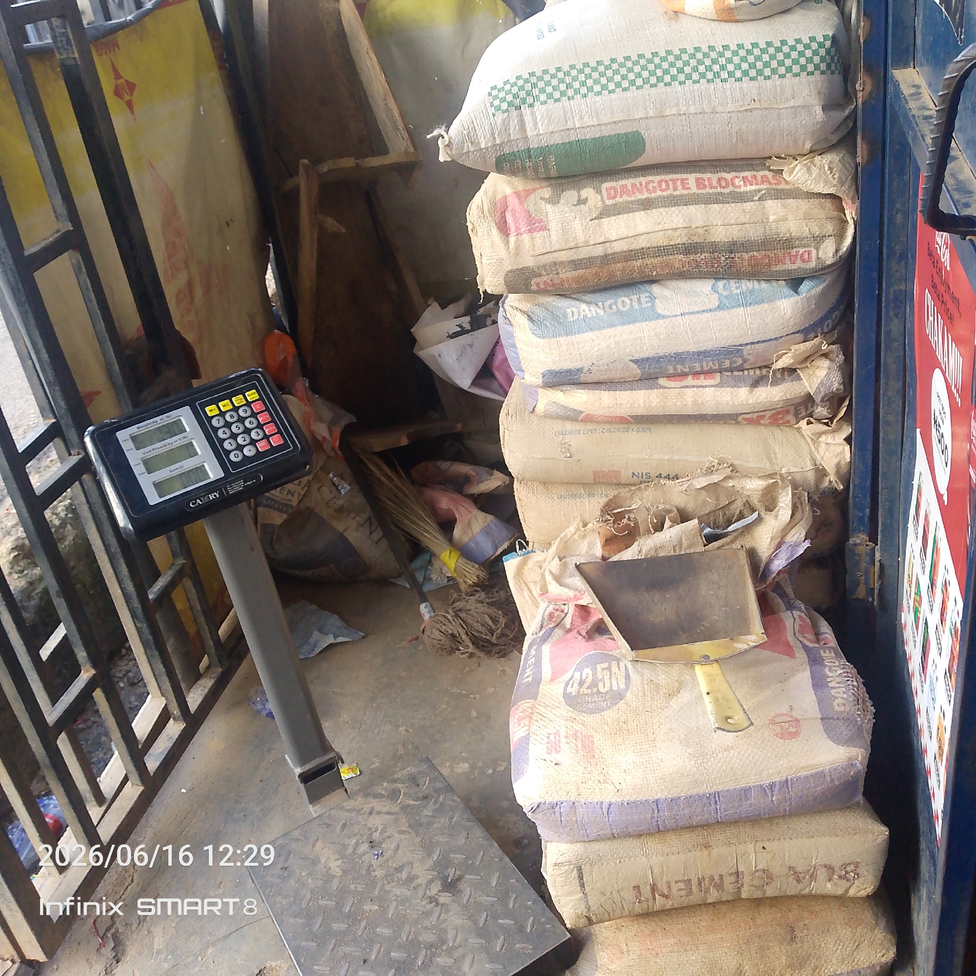 In an outdoor urban setting, a stack of cement bags is piled next to a metal gate. A digital scale, with the brand name "CAMRY" visible, stands to the left of the cement bags, its display screen showing numerical data. The bags are made of woven material and feature various brand names and markings, including "DANGOTE BLOCKMASTER" and "42.5N GRADE CEMENT." Some of the bags are stacked precariously, with one bag at the top having a yellow metal tool resting on it. A broom and some coiled rope are also visible on the ground near the bags. The scene is illuminated by natural light, and the ground is a concrete or paved surface. A red sign with text and graphics is attached to the blue metal gate on the right side of the image. A timestamp in the lower left corner indicates "2026/06/16 12:29" and the device "Infinix SMART 8".