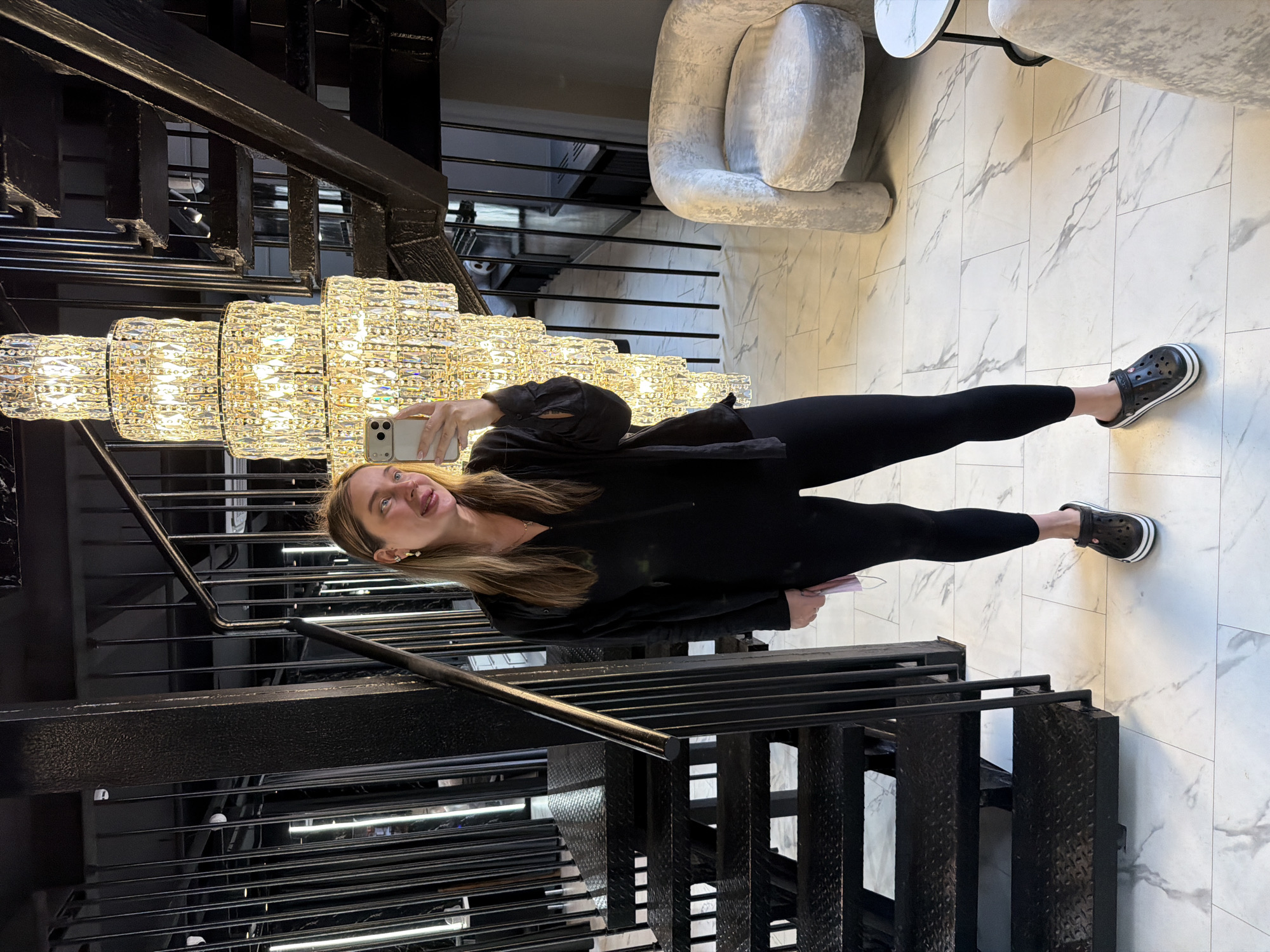 The image shows a woman taking a selfie while standing on a staircase. The staircase is made of black metal and has multiple levels. A large, ornate chandelier hangs nearby. The woman is wearing black leggings, a black top, and black Crocs. She is holding a phone in her hand and sticking out her tongue. The background consists of a white marble floor and a modern chair. It is possible that this image was taken in Moscow, Russia, but there is no explicit information confirming this.