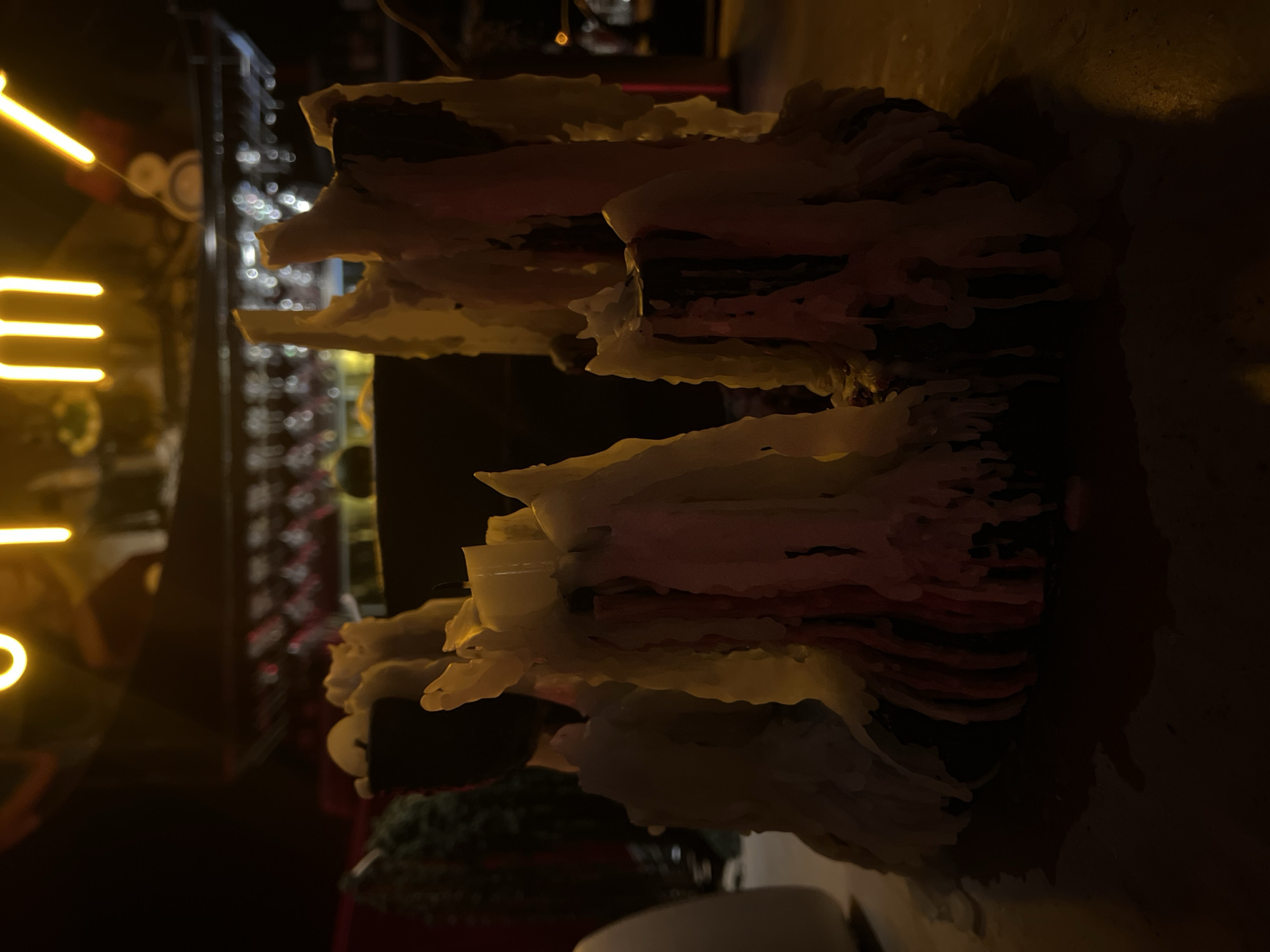The image shows a heavily layered candle, predominantly white and red, with significant wax buildup. The background is blurred and dark, but there are hints of a more brightly lit area with linear yellow lights and what appears to be shelves with various items, possibly bottles. The overall scene is dimly lit, casting shadows on the candle and surrounding area. The location of the image, specifically Mengwi, Indonesia, does not provide enough information to establish a broader context without additional details.