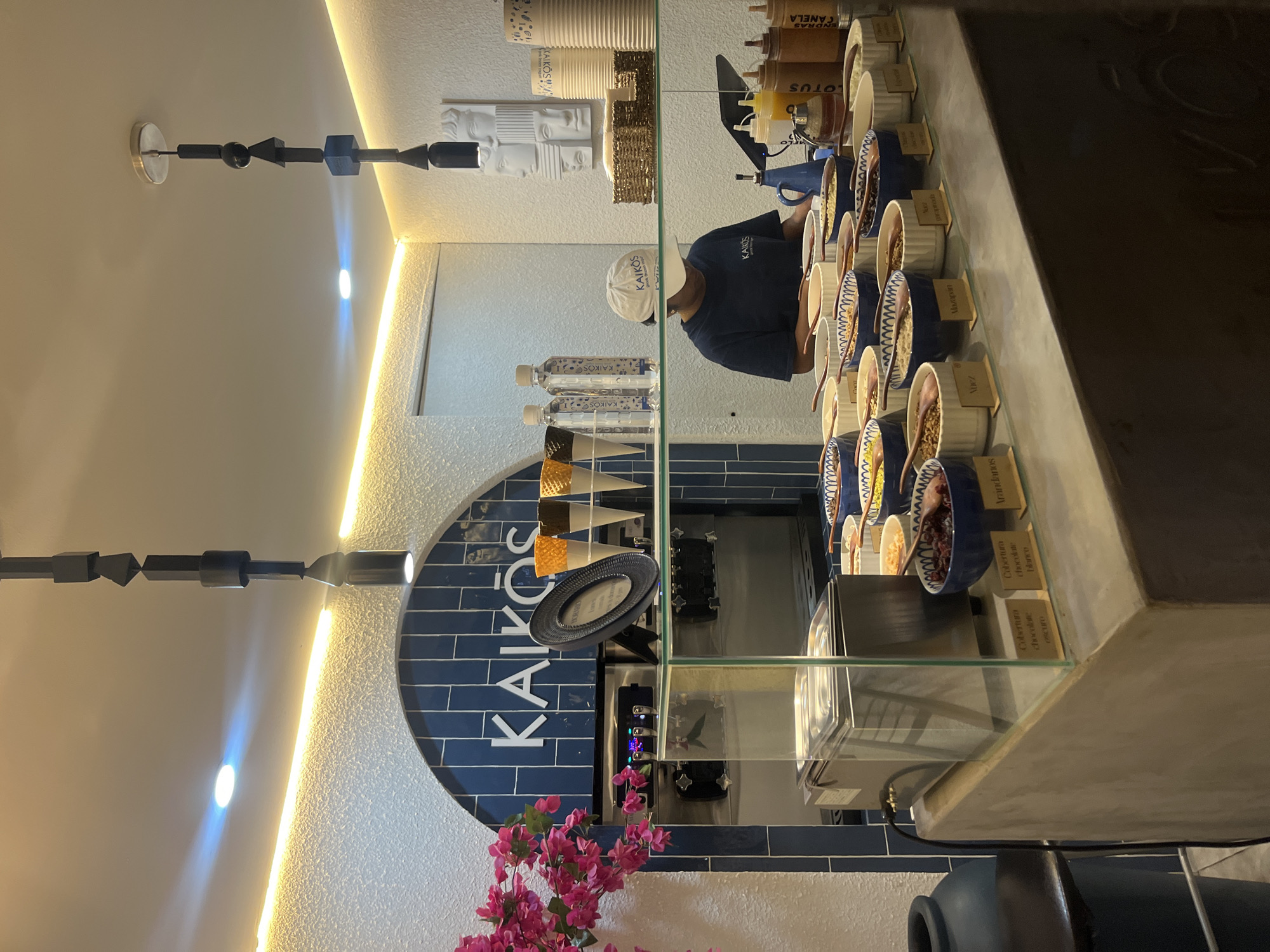 Here's a description of the image:
The image shows the interior of a "Kairos" ice cream shop. A person wearing a blue shirt and a Kairos branded hat stands behind the counter, presumably preparing or serving ice cream. The name of the shop, "KAIKOS" is prominently displayed on a blue tiled wall in the background. Above the counter are rows of topping containers. Below the toppings is the ice cream freezer and on the counter are written labels for the ingredients. The surrounding decor includes a light fixture comprised of geometric shapes, and decorations such as a set of faces in white and pink flowers. The presence of Spanish language labels ("Cobertura Chocolate Blanco/Oscuro") suggests the location is in a Spanish-speaking area. The shop is located in Puebla, Mexico.