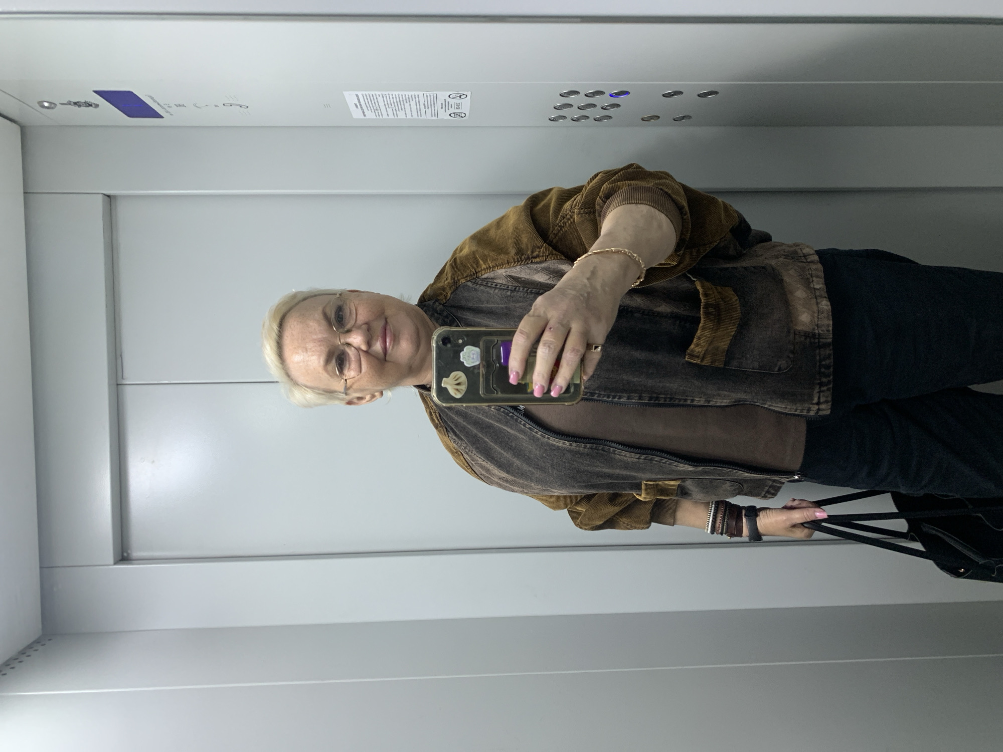 A Caucasian woman with short white hair, eyeglasses, and pink painted fingernails is taking a selfie in what appears to be an elevator. She is wearing a brown t-shirt, a brown corduroy jacket, and dark denim pants. She is holding a smartphone with stickers on the back in her right hand, and a black bag in her left hand. The elevator has light gray walls, a panel with buttons, and a sign in Russian. The location is likely Chaikovsky, Russia, based on the language on the sign.