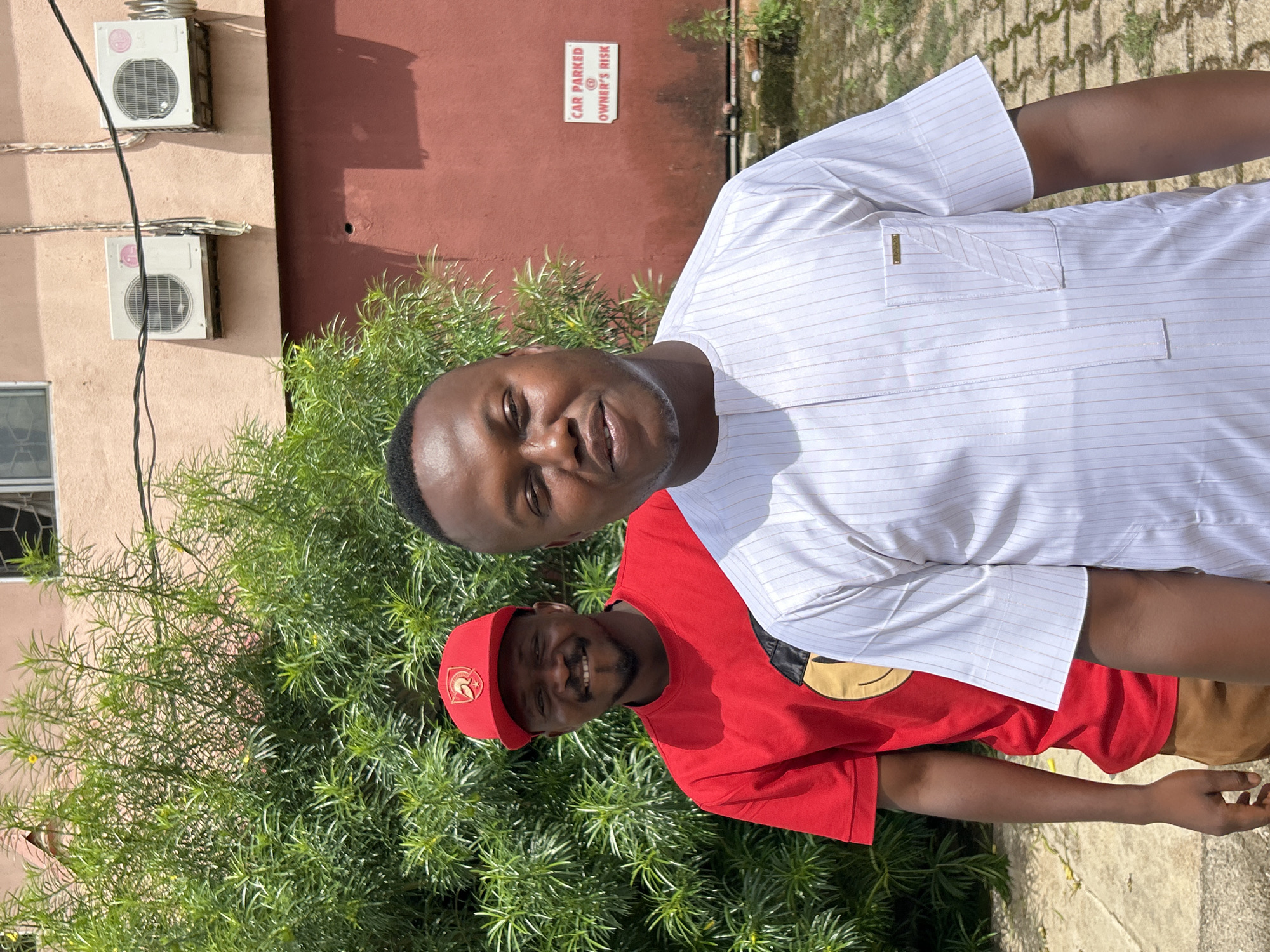 This image depicts two dark-skinned males standing outdoors in Jos, Nigeria, captured during daylight hours.
The man positioned closer to the camera, on the right, is wearing a short-sleeved white top with thin vertical brown stripes. The top features a distinct left breast pocket with diagonal stitching and a small rectangular gold-colored metal tag. He has a closely cropped hairstyle and a light beard, looking slightly to the viewer's right with a neutral expression.
Behind and to the left of the first man, the second man is visible, wearing a red short-sleeved t-shirt with a yellow and black "smiley face" graphic on the chest. He wears a red baseball cap adorned with a gold-colored emblem, appearing to be a stylized helmet, on the front. This man has a beard and mustache, smiling broadly directly at the camera.
The background consists of dense, vibrant green foliage with small yellow blossoms, partially obscuring a building. The upper section of the building is painted a light salmon or reddish-brown hue, featuring a window on the left side and two white outdoor LG air conditioning units mounted on the wall with visible wiring. Below this section, the building transitions to a darker red-brown color. A white rectangular sign with red lettering, stating "CAR PARKED @ OWNER'S RISK," is affixed to this darker wall. The ground in the foreground is paved with light-colored, interlocking stones.