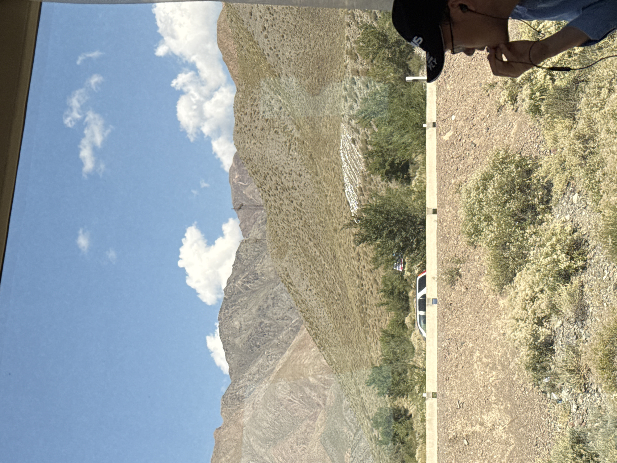 The image shows a view from a vehicle, looking out at a mountainous landscape. The sky is blue with scattered white clouds. In the foreground, a person wearing a blue shirt and a black baseball cap with a logo is visible on the right side of the frame, facing away from the camera and towards the landscape. They are wearing earbuds and appear to be touching their face with their right hand. A concrete barrier runs horizontally across the middle of the frame, separating the foreground from the background. Beyond the barrier, there are green trees and a white SUV. In the distance, rugged mountains with dry vegetation cover their slopes are visible, with a power transmission tower on one of the peaks. The scene depicts a person observing a natural, semi-arid environment. This image is not taken in Kok Moynok Eki, Kyrgyzstan.
