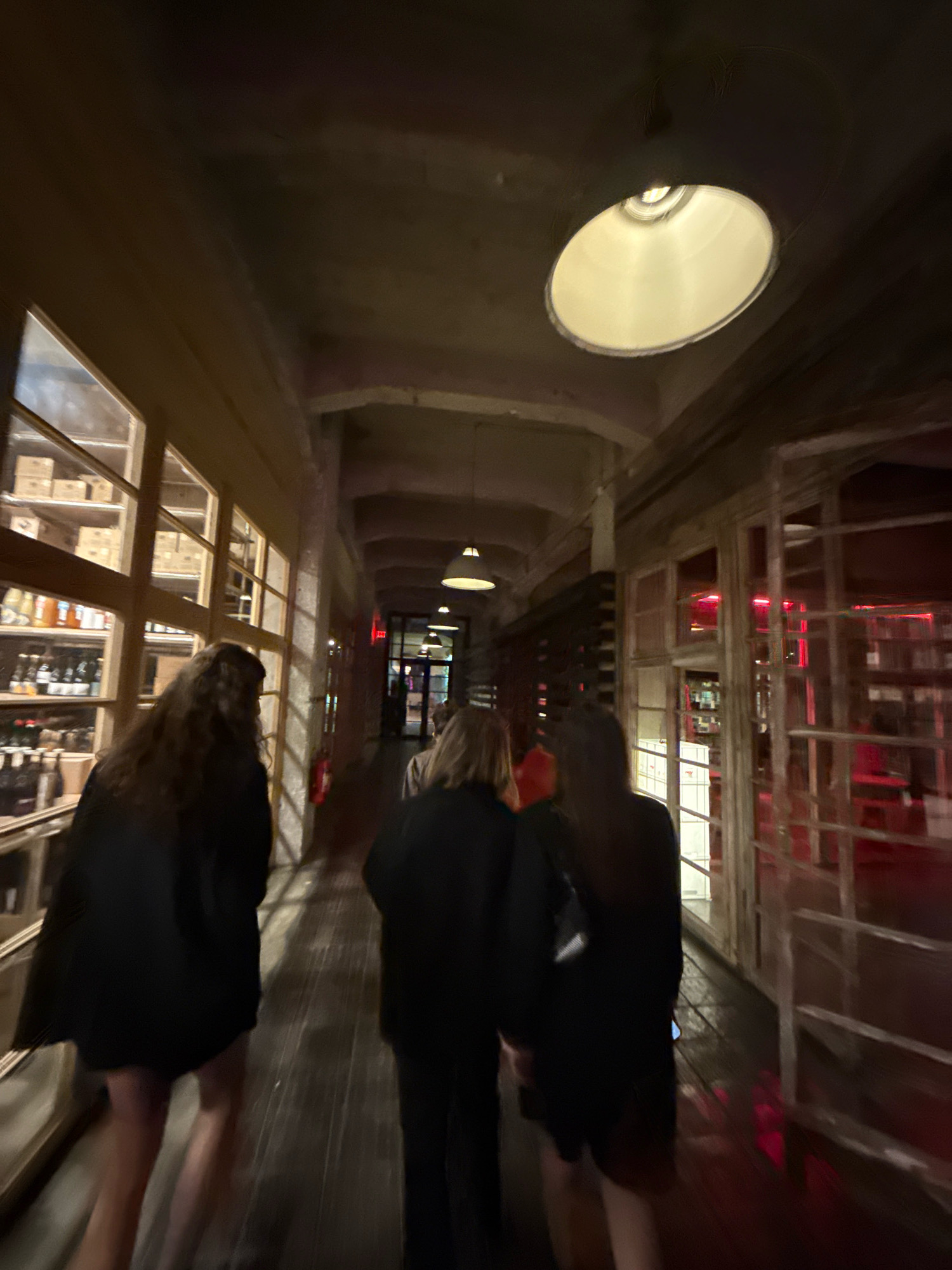The image depicts an interior view of a dimly lit establishment, possibly a bar or restaurant, with wooden floors and exposed concrete ceilings. Three individuals, two women and one person of indeterminate gender, are walking away from the camera down a corridor. The woman on the left has long, dark curly hair and is wearing a black outfit. The person in the middle is wearing a dark outfit and has blonde hair. The woman on the right is wearing a black outfit and also has blonde hair. Large windows line the left side of the corridor, revealing shelves stocked with bottles and boxes. The right side of the corridor features more windows, behind which red lighting is visible, suggesting another room or area. Industrial-style pendant lights hang from the ceiling. The overall atmosphere is moody and atmospheric, consistent with a nighttime scene in an urban environment. The presence of architectural elements and the specific lighting suggest a location in Tbilisi, Georgia, known for its industrial-chic venues.