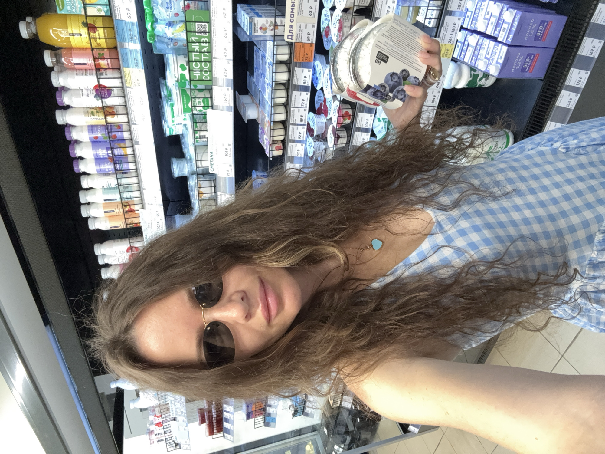 A woman with long, brown, wavy hair and dark sunglasses is taking a selfie in a supermarket aisle. She is wearing a light blue and white checkered dress and a silver necklace with a turquoise heart pendant. In her right hand, she is holding a container of blueberries and yogurt. The background shows shelves stocked with various dairy products, including yogurt, milk, and sour cream. The pricing tags on the shelves are visible. The environment appears to be a well-lit supermarket. The text on some of the products and price tags indicates the location is likely in Russia, given the Cyrillic script and currency symbol "P" for Rubles.