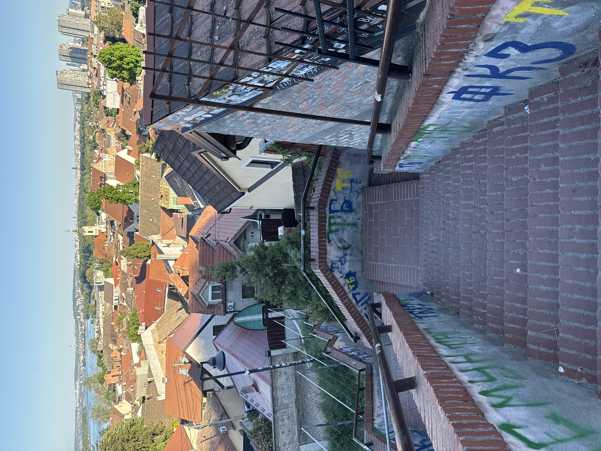 The image shows an elevated view of Zemun, a municipality in Belgrade, Serbia. The perspective is from a high point overlooking the city, capturing red-roofed buildings, the Danube river in the distance, and modern high-rise buildings further in the background. In the foreground, there are brick steps with graffiti, leading downwards, and metal railings alongside the steps. The lighting suggests it is daytime.