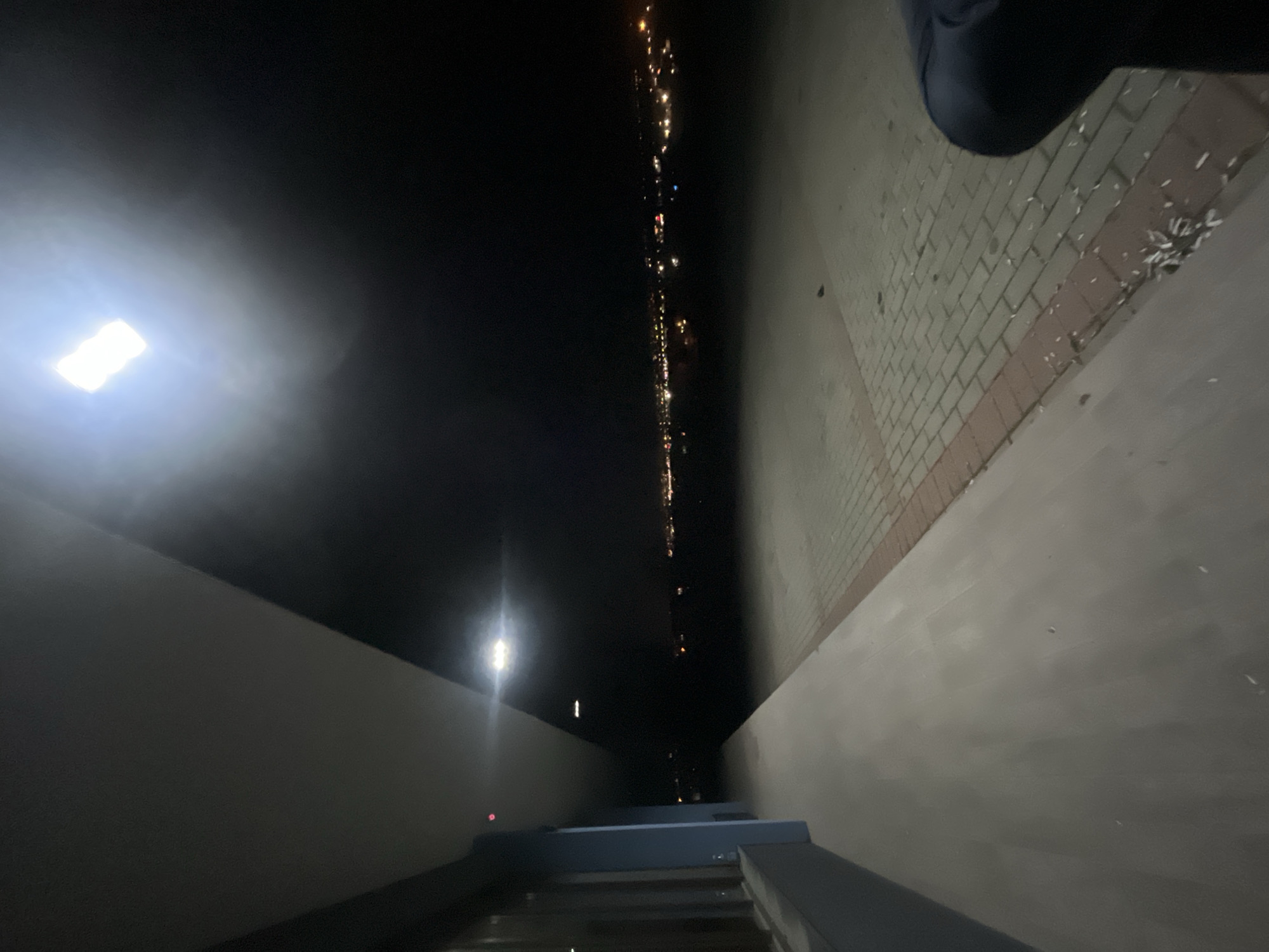 A night shot taken at an angle shows a staircase leading down, flanked by walls on either side. To the left, a bright, square-shaped light source illuminates the wall. In the distance, a line of blurred lights indicates the presence of a city or town; this is Asenovgrad, Bulgaria. The wall on the right has a tiled surface. The staircase appears to be wet. A dark shape is visible in the upper right corner, potentially a person or object in the foreground.