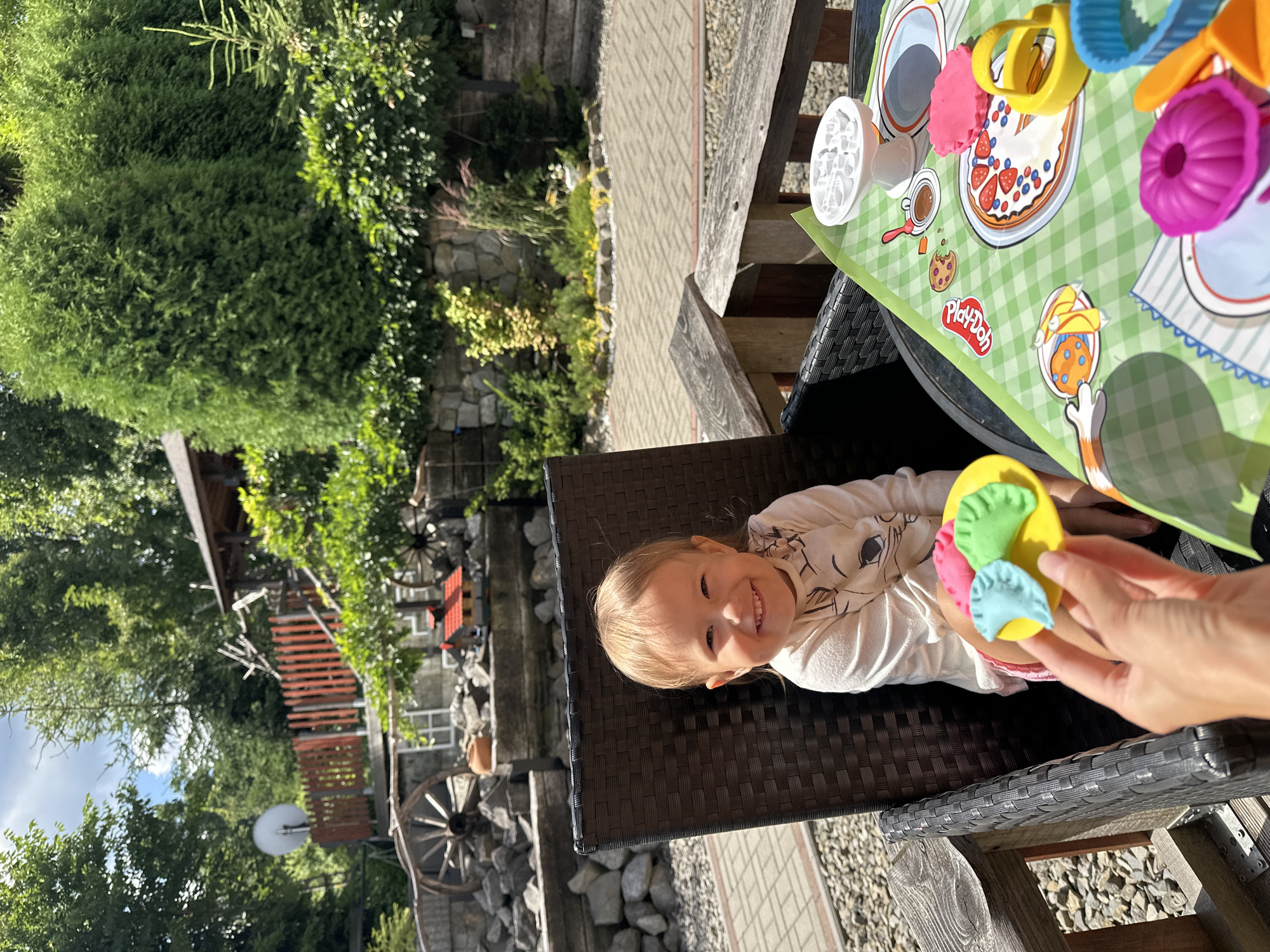 A young girl with blonde hair and a light-colored shirt sits in a black wicker chair outdoors. She is smiling broadly. In front of her is a Play-Doh mat with various food-themed molds and accessories. A hand is holding a yellow plate with Play-Doh shapes resembling pierogi. The background shows a stone wall, greenery, trees, and a rustic wooden structure. The location appears to be a backyard or patio in Wisła, Poland, a city known for its mountainous terrain and wooden architecture.