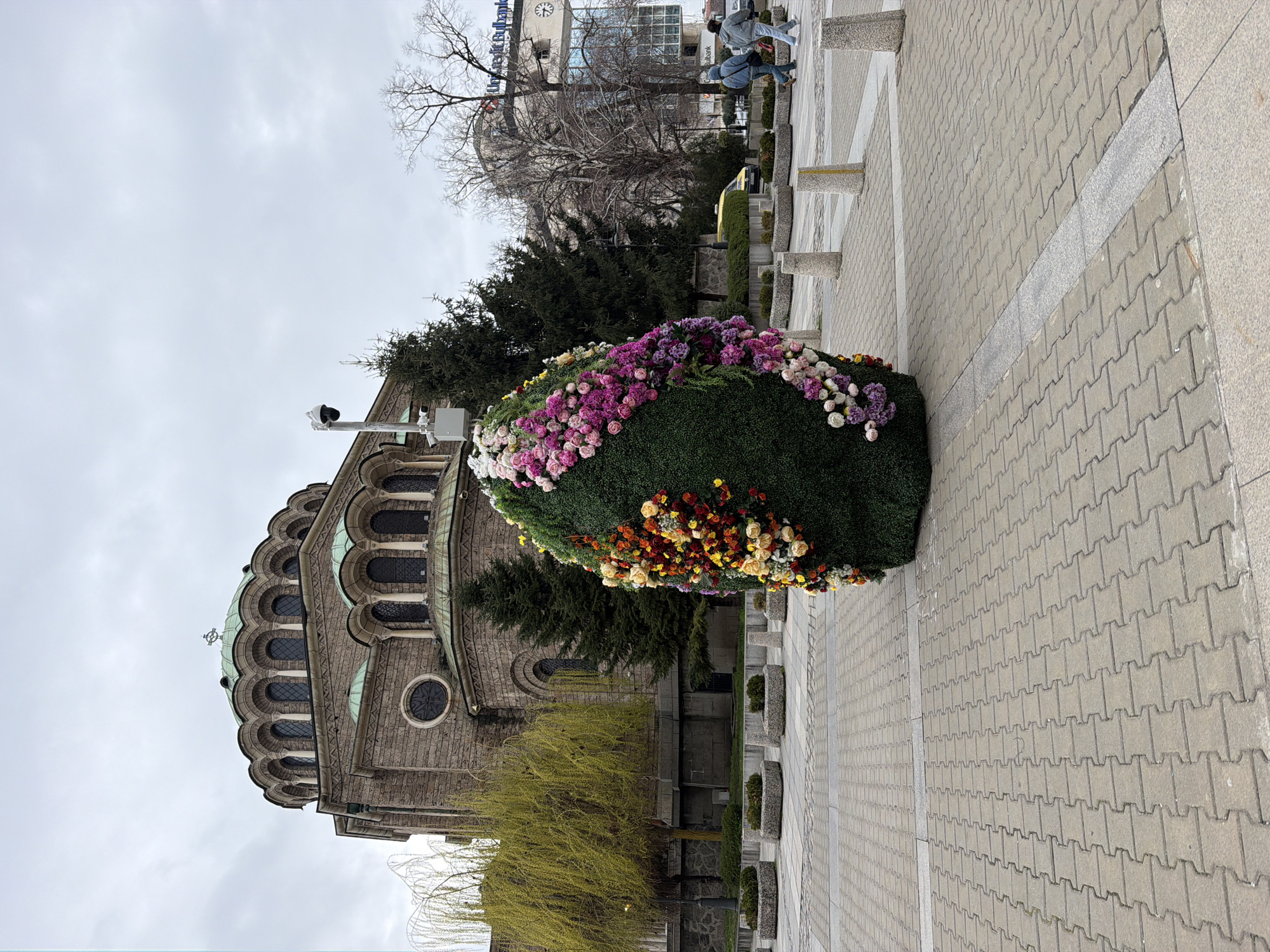This photograph depicts a street scene in Sofia, Bulgaria. In the foreground, a large, ornate Easter egg made of greenery and colorful flowers serves as a decorative centerpiece. The egg is positioned on a paved walkway made of interlocking blocks.
To the left of the floral display is the Saint Alexander Nevsky Cathedral, a prominent landmark in Sofia. The cathedral's architectural details, including its arched windows and the green dome, are visible. A willow tree, with its branches displaying fresh spring foliage, is located near the cathedral.
In the background, across the street, is a clock tower atop a commercial building. Two individuals are visible on the sidewalk in the distance, walking past the clock tower. The sky is overcast.