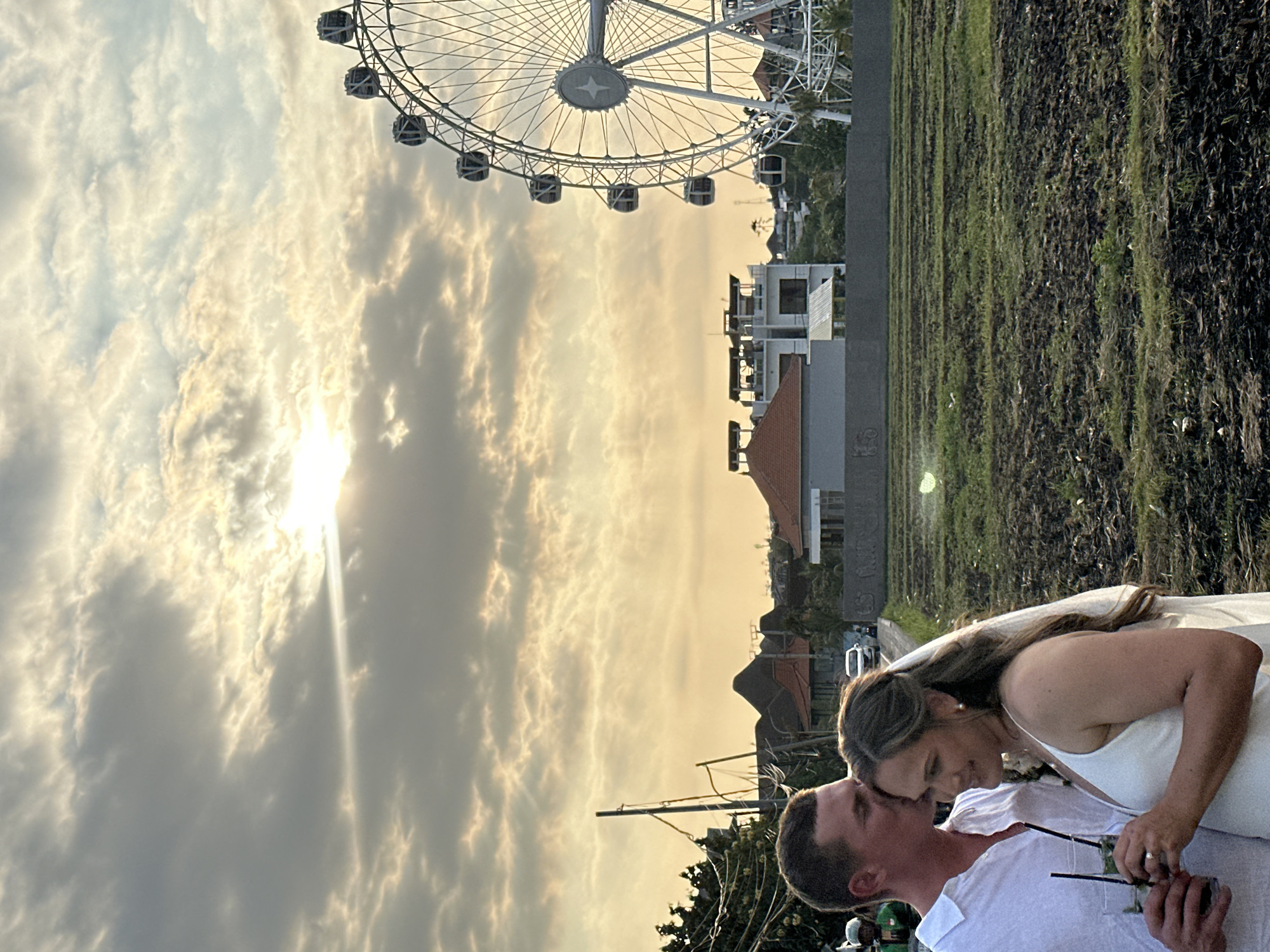 A man in a white shirt kisses a woman in a white wedding dress on the forehead. They are holding drinks. The woman is wearing a veil and pearl earrings. In the background, a Ferris wheel stands against a cloudy sky with the sun breaking through. Buildings and trees are visible between the couple and the Ferris wheel. The ground in front of the couple is a tilled field. The scene appears to be located in North Kuta, Indonesia, likely near a tourist attraction.
