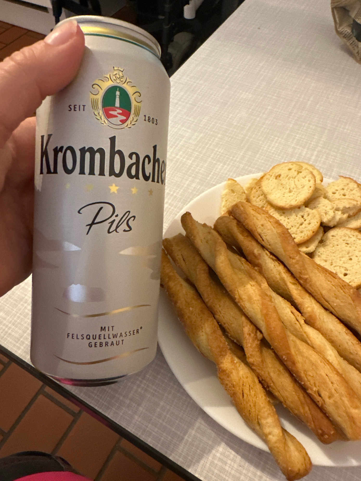 A hand holds a can of Krombacher Pils beer, which is silver with a gold and green logo. Next to the can on a white plate are several breadsticks and small, round crackers. The scene is set on a grey, textured tablecloth. The background is blurred but suggests an indoor setting with a hint of a tiled surface and a brown object. The image appears to have been taken in Krefeld, Germany.