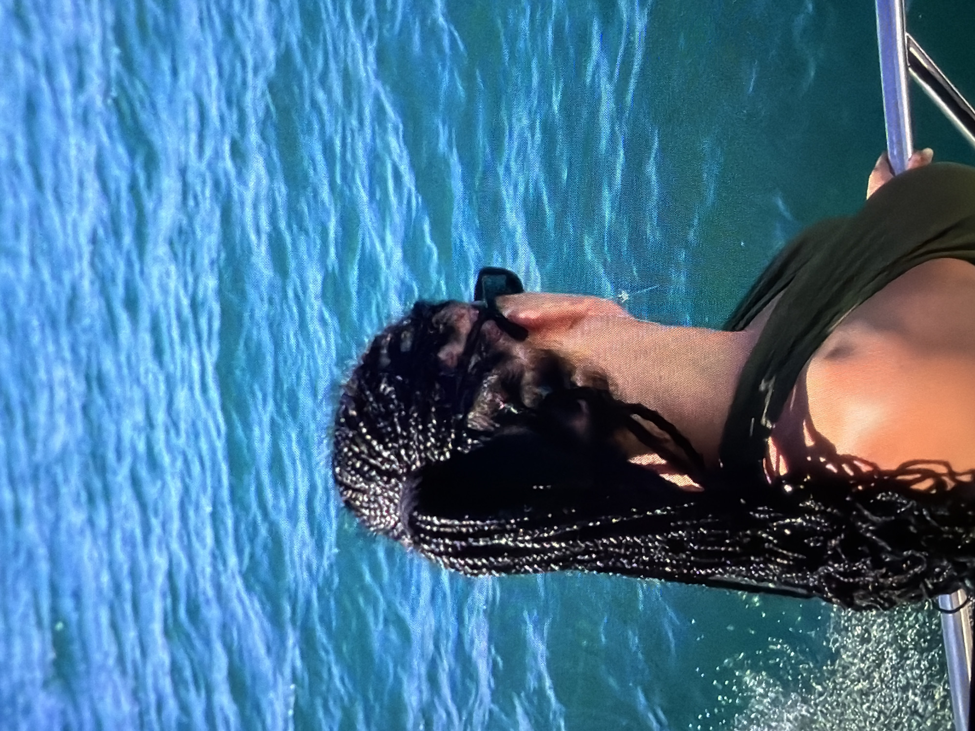 A woman with braided hair and dark sunglasses sits on a boat, with blue water visible in the background. She is wearing a dark green top. The boat has a metallic railing. The location is unknown.