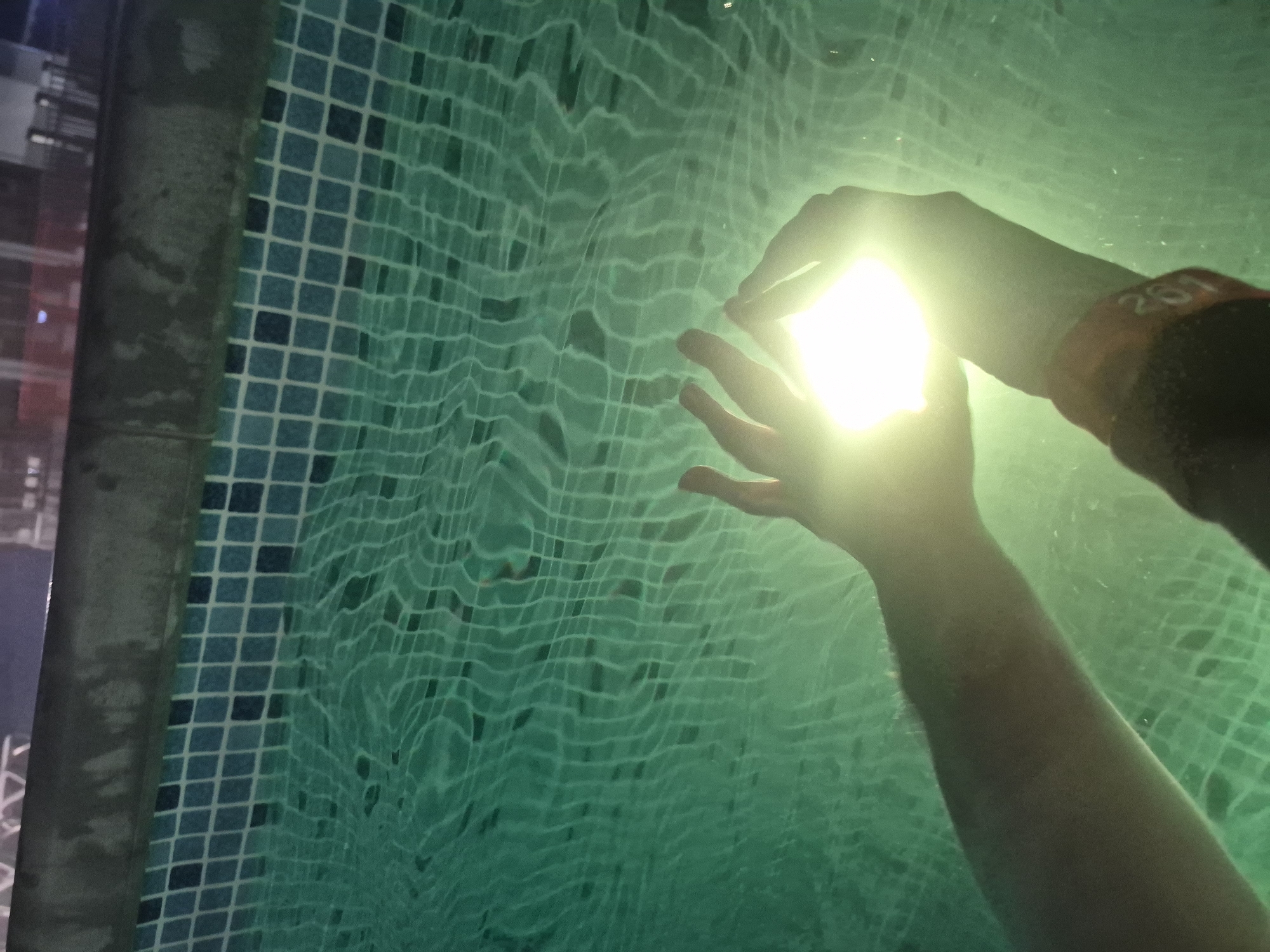 The image presents a close-up, top-down view of two hands submerged in a swimming pool, likely at night or in a dimly lit indoor environment. A very bright, square-shaped light source emits intense white light from between the hands, casting a strong glow and causing a lens flare effect. The hands appear to be holding or interacting with this radiant object underwater.
The water itself is a translucent greenish-blue, displaying pronounced ripples and distortions on its surface that refract the light. Below the water, the pool's bottom and an adjacent wall are visible, tiled with a grid pattern of small blue and green squares. The water's movement distorts the appearance of these tiles.
On the wrist of the right hand, an orange band is visible, clearly displaying the numbers "261". To the far left of the frame, a dark, blurry vertical structure, possibly a pillar or the edge of the pool deck, stands next to the tiled wall. Beyond this, the background is very dark with faint, out-of-focus lights, suggesting an urban setting or building lights in the distance. The overall scene emphasizes the interaction with the luminous object in the water, set against the backdrop of the tiled pool. The location context is Votkinsk, Russia.