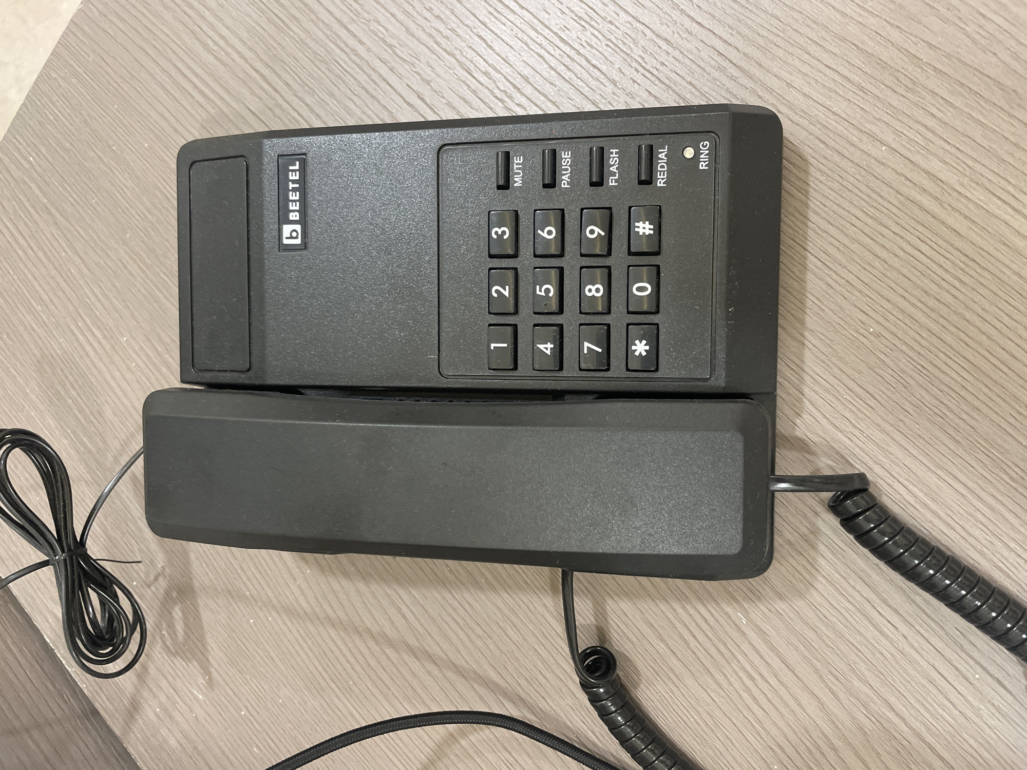 The image shows a black Beetel brand telephone on a light wooden surface. The phone consists of a rectangular base with push-button numbers and function keys (Mute, Pause, Flash, Redial, Ring) arranged in a 3x4 grid. The logo "b BEETEL" is visible on the left side of the base. The handset rests on top of the base. A coiled cord connects the handset to the base. Another cord connects the base to an external power source. There is a small bundle of the phone wire on the side next to the base. The lighting is relatively even. The location is unspecified.