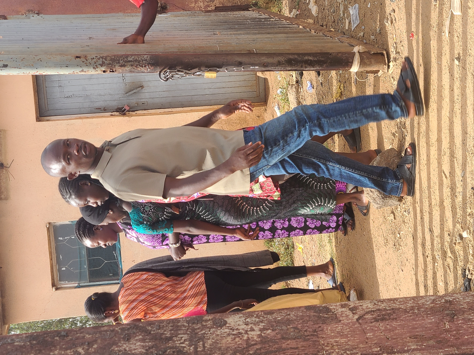 A group of four individuals, comprising three women and one man, stand adjacent to a building in an outdoor setting. The man is positioned at the front of the group, wearing a beige short-sleeved shirt and blue jeans, with dark sandals on his feet. The women stand behind him. The woman closest to the man is wearing a patterned dress of purple and black with a green top. A window is visible in the peach-colored wall of the building. The ground is covered in a combination of sand and detritus. A metal gate, secured with a chain and padlock, is visible on the wall. The scene is illuminated by natural daylight. The location is Jos, Nigeria.