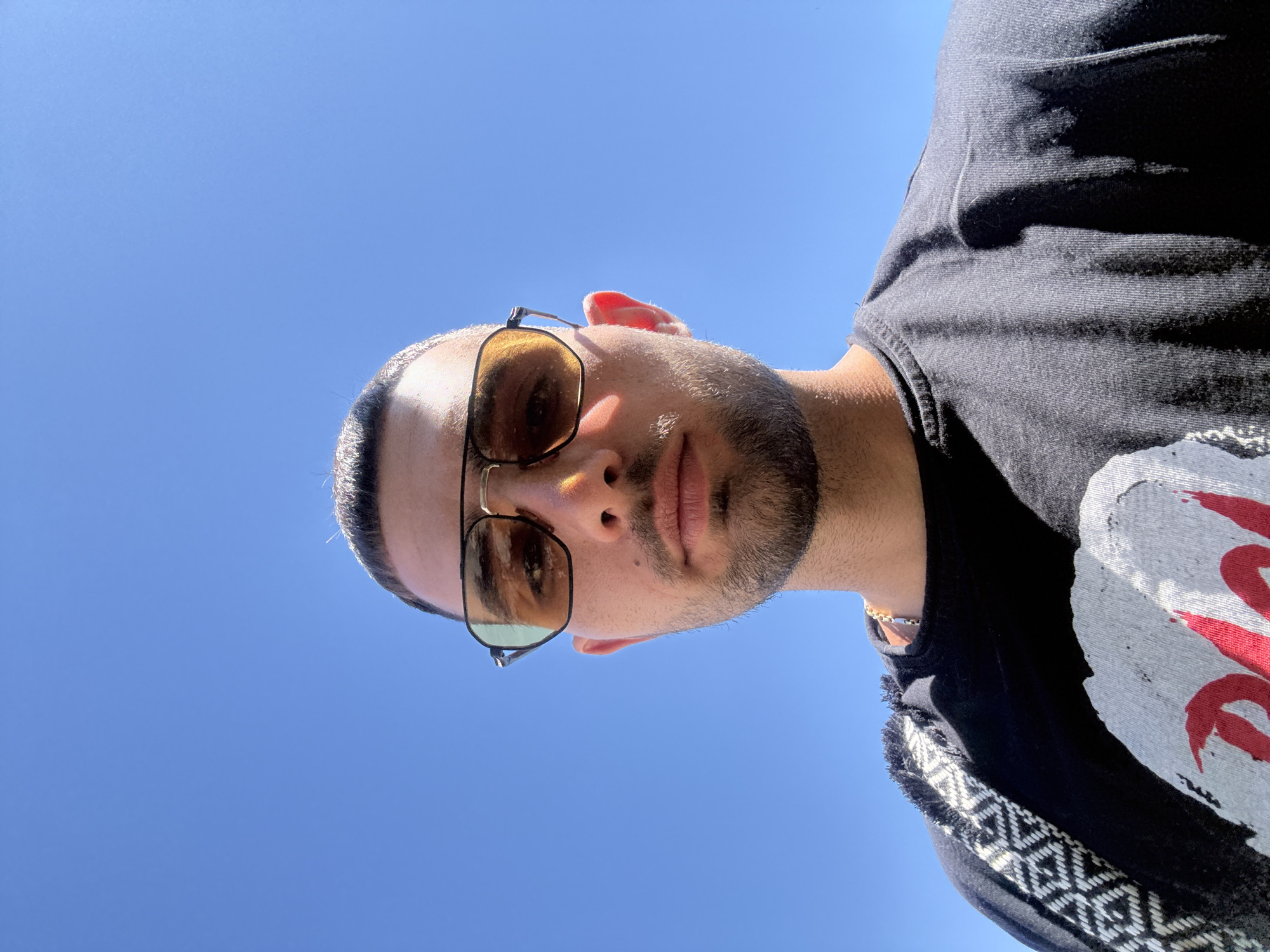 A low-angle, head-and-shoulders selfie of a young man with short hair and a goatee is captured against a clear, bright blue sky. He is wearing sunglasses with yellow lenses and a black t-shirt with a graphic design on the front. A portion of a patterned bag strap is visible over his left shoulder. The image appears to have been taken outdoors in Nesebar, Bulgaria.