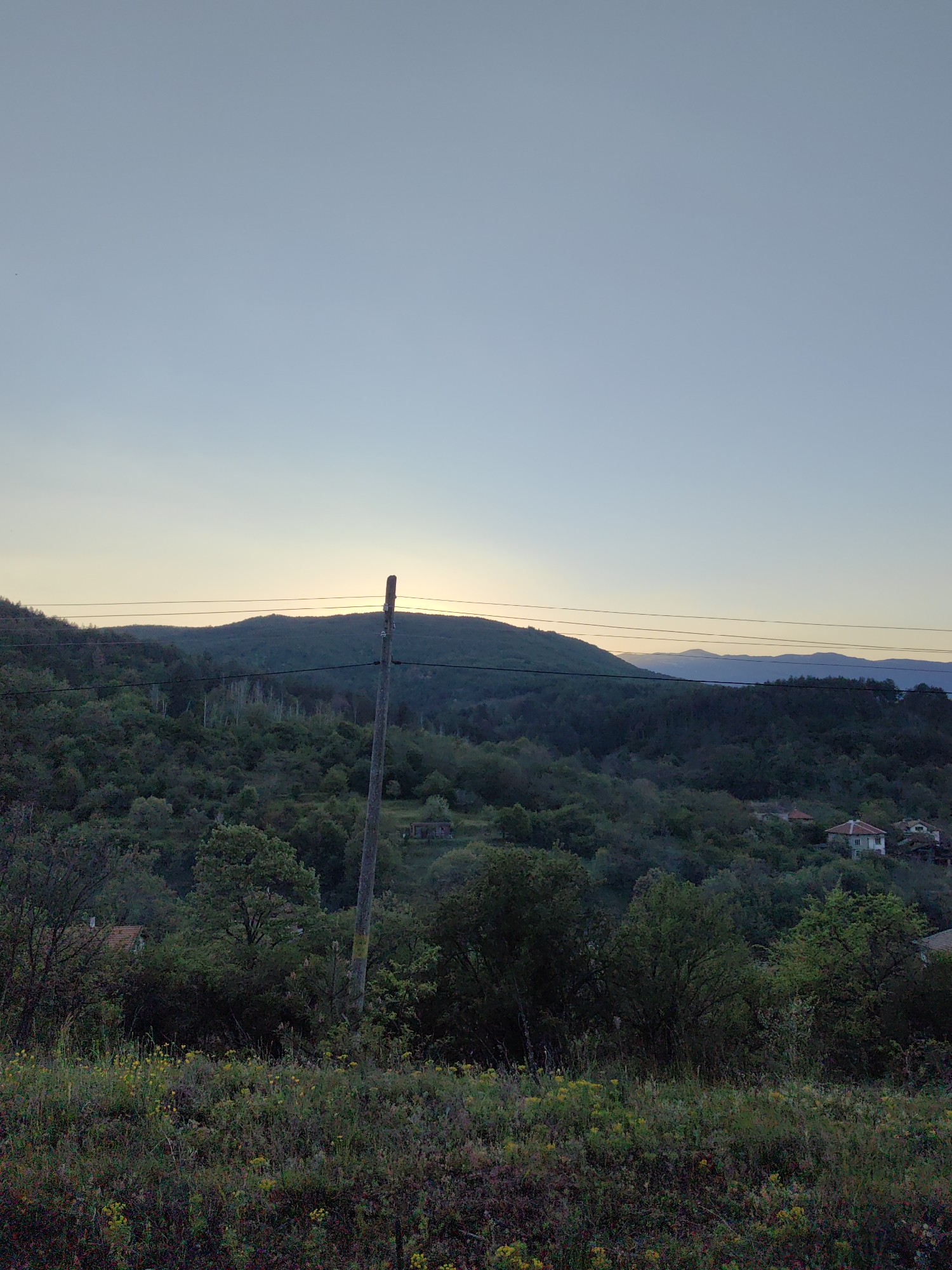 The image presents a scenic view from Hristo Danovo, Bulgaria. A wooden utility pole stands in the foreground, with power lines stretching horizontally across the frame. Behind the pole is a heavily wooded hillside. In the background, a distant mountain range is visible. In the mid-ground some houses are visible. The sky is mostly clear, with a subtle color gradient suggesting either sunrise or sunset. A foreground of tall vegetation is visible at the bottom of the frame.