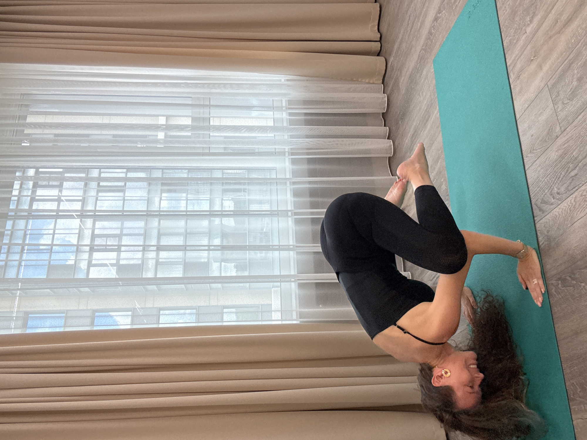 A woman is performing a yoga pose on a teal mat in a room. She is wearing a black leotard and leggings, with her hair curly and long. The room has wooden floors and is lit by natural light coming through a large window covered by beige curtains and sheer white curtains. The building across the street is visible through the window. The scene appears to be in the city of Khimki, Russia.