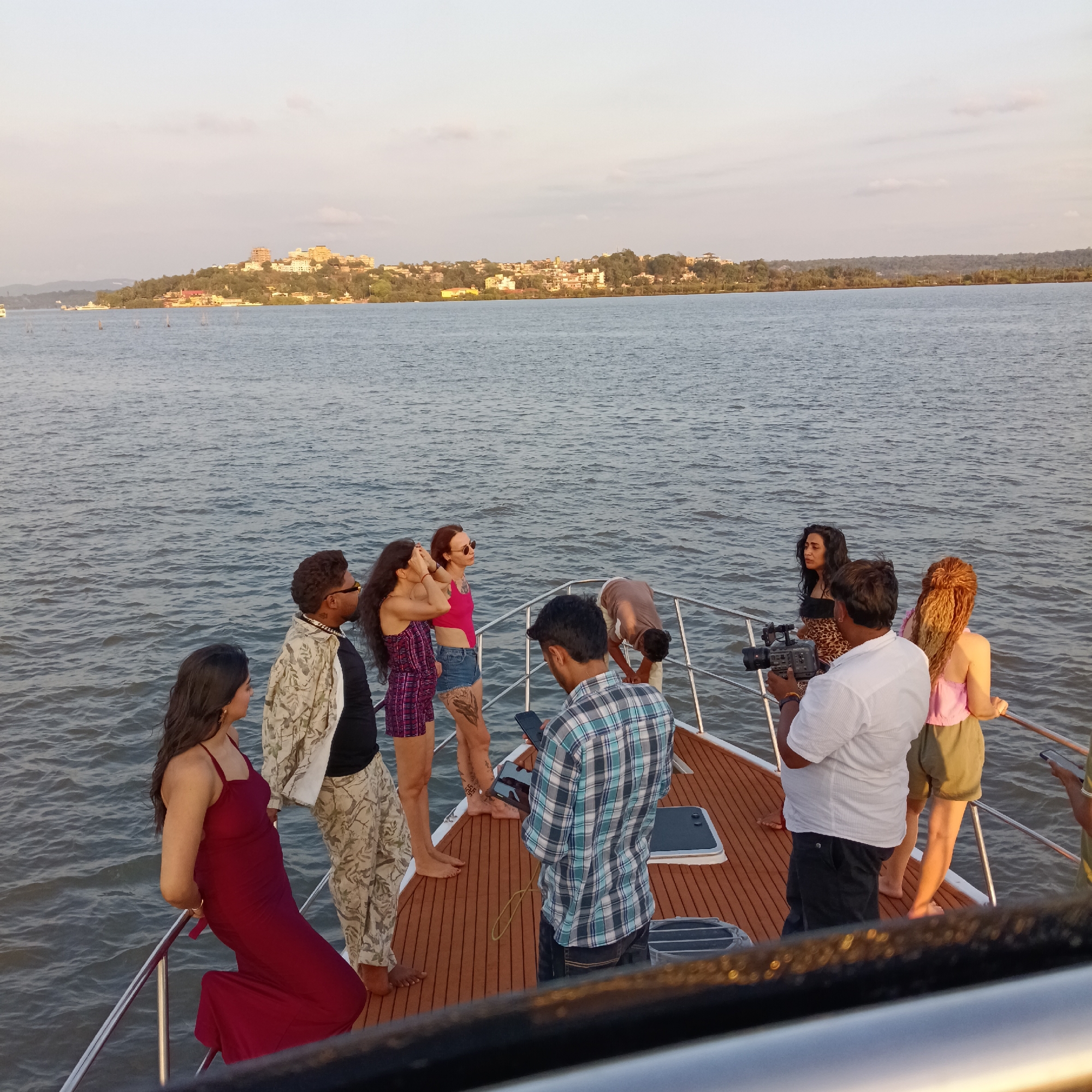 Captured on a boat navigating waters near Britona, India, the image portrays a group of people engaged in what appears to be a photoshoot or filming session. Three women stand towards the front of the boat, wearing casual summer attire. One is in a red dress, another in a red top and denim shorts, and the third in a checkered romper. A man in a printed shirt and another man in a plaid shirt stand nearby, both holding cell phones. Further back, a man with a camera films two women, one with dark hair and the other with red/blonde hair. In the background, a landscape of buildings and greenery is visible, likely situated on the banks of the water. The sky is lightly overcast, indicating an overcast day.