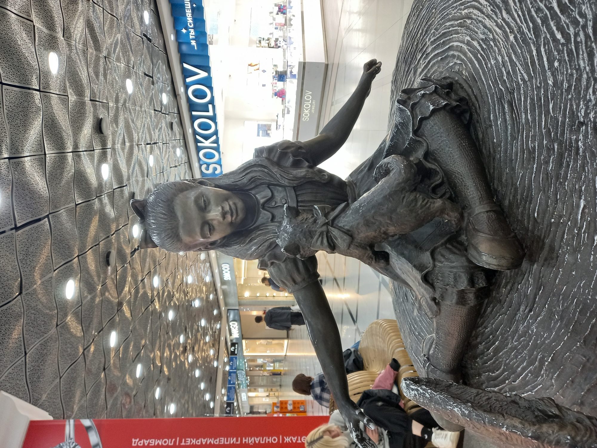 A bronze sculpture of Alice, from Alice in Wonderland, is displayed in a shopping mall in Yekaterinburg, Russia. Alice is depicted sitting with a cat in her lap. The sculpture is placed on a circular base that resembles rippling water. In the background, there are storefronts for various shops, including Sokolov, Mango, and S Mood. Several people are visible in the mall, including shoppers browsing the stores and sitting on benches. The ceiling of the mall is made of reflective panels with embedded lights, creating a textured and illuminated effect. A red pillar with Russian text is visible on the left side of the frame.