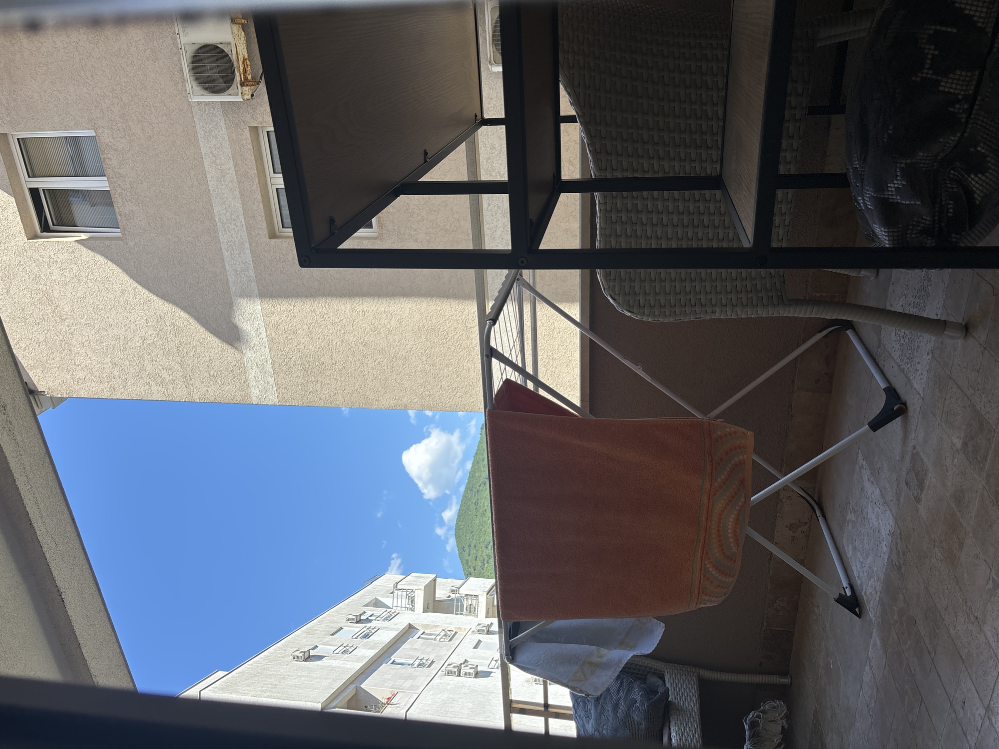 The image depicts an upward view from a balcony in Budva, Montenegro. The foreground is dominated by balcony furniture, including a table and chair set with a woven design, and a clothes-drying rack. An orange towel and other articles of clothing hang on the rack. In the background, a clear blue sky is visible, framed by the surrounding buildings. One building appears to be a modern apartment complex with multiple windows and balconies. In the distance, a green, mountainous landscape is seen through the opening. The view suggests a residential area within the city of Budva, with a mix of urban and natural elements.