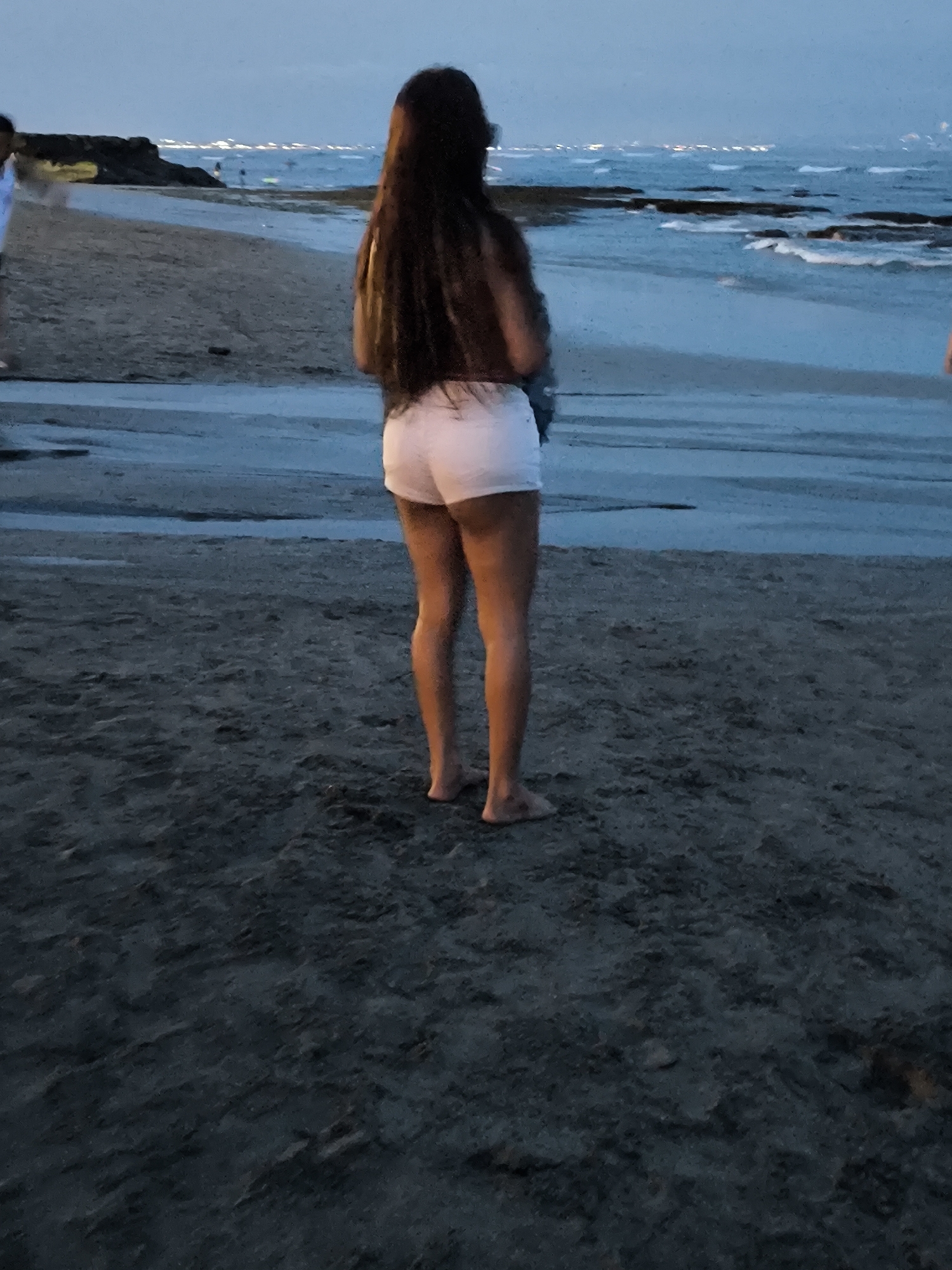 A woman with long, dark hair stands on the beach at Canggu, Indonesia. She faces the ocean, wearing white shorts. The sand is dark and wet, indicating a recent tide or wet conditions. In the background, the ocean is visible with waves breaking near the shore, and in the distance, faint lights suggest the coastline. Other people are visible in the background.