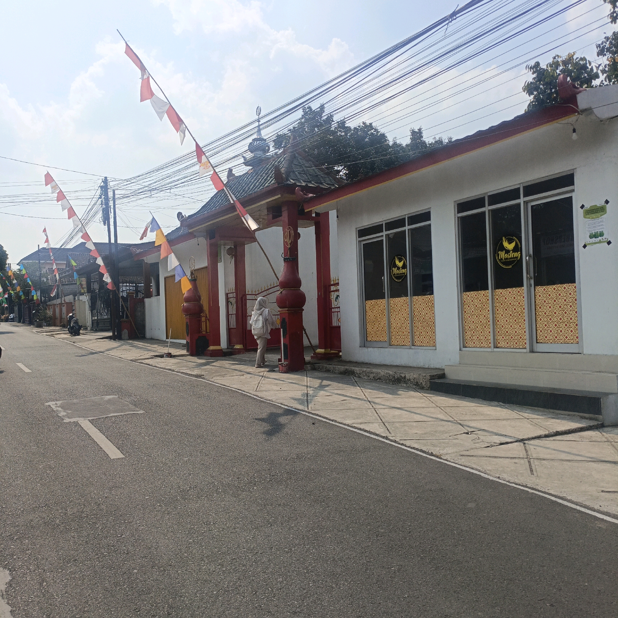 The image depicts a street scene in Salatiga, Indonesia, decorated with Indonesian flags, which are red and white. The street is paved, with a white line marking the edge. A sidewalk runs alongside the street, made of concrete tiles. Buildings line the sidewalk, one with a white facade and large glass windows featuring a golden geometric pattern. Another building, possibly a mosque or temple, has a traditional tiled roof and red pillars. A person wearing a light-colored hijab and backpack is walking on the sidewalk. Several motorcycles are parked along the street and sidewalk. Overhead, a dense network of electrical wires crisscrosses the sky, supported by utility poles. The sky is partly cloudy, with the sun casting shadows on the ground, indicating it is daytime.