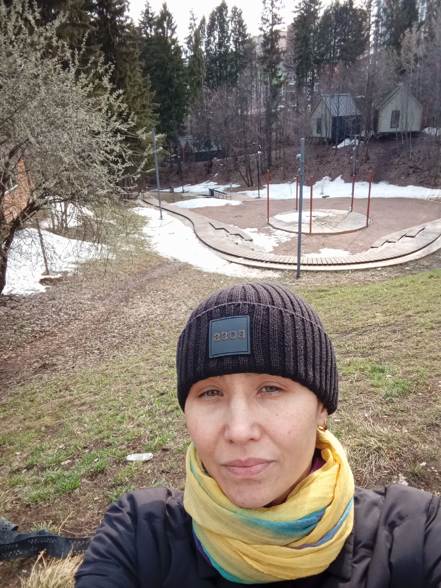 The image is a selfie of a woman wearing a dark-colored beanie with the word "BOSS" on a square patch, a dark jacket, and a yellow scarf with blue accents. She is positioned in the foreground.
Behind her is a park setting with melting snow. A circular playground area with red poles is visible. In the background, there are bare trees, evergreen trees, and two small houses or cabins on a hillside.
Based on the presence of these distinctive cabins and playground, the location is likely near the Sosnovy Bor (Pine Forest) Park in Izhevsk, Russia. The season appears to be early spring, indicated by the melting snow and bare trees.