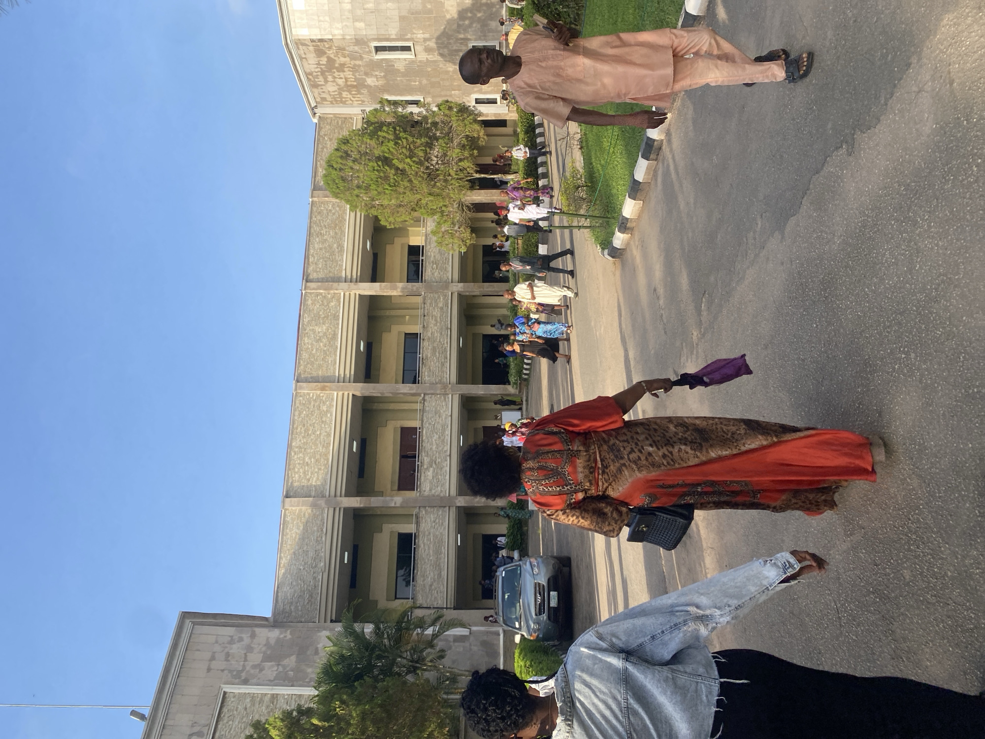 The image depicts a street scene in Ota, Nigeria. In the foreground, a woman in an orange and brown patterned dress walks holding a purple item, possibly a handkerchief. Beside her, partially visible, is a person wearing a denim jacket and black pants. In the background, a group of people is gathered in front of a multi-story building with concrete architecture. A parked silver car is visible in the middle ground. Another man, dressed in peach-colored clothing, is walking on the right side of the image. The street is paved, and the sky is clear and blue.