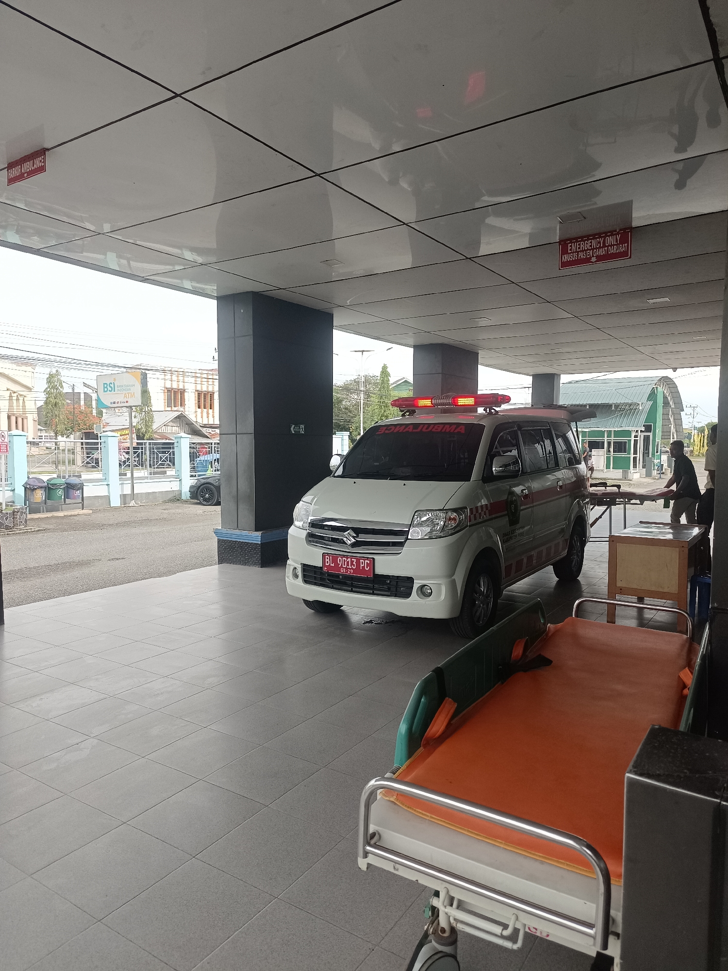 The image depicts an outdoor scene under a covered walkway, likely at a medical facility, given the presence of an ambulance and stretchers. A white ambulance with its emergency lights activated is parked in the foreground. The ambulance has the word "AMBULANCE" written on its side in reverse and a license plate that reads "BL 9013 PC". Two stretchers are visible; one is orange with metal railings and wheels, positioned in the foreground, and another is partially obscured behind a wooden table in the background.
In the background, beyond the covered walkway, a street is visible with buildings and trees. A sign for "BSI" (Bank Syariah Indonesia) with an ATM is also present, indicating a commercial area near the facility. Two individuals are seen near the stretcher in the background, one appearing to be positioning the stretcher. There are also signs on the ceiling of the walkway: one reads "PARKING AMBULANCE" and another reads "EMERGENCY ONLY KRUSUS PASIEN GAWAT DARURAT". The overall environment suggests a functional, perhaps busy, area related to patient transport and medical services. The lighting indicates it is daytime.