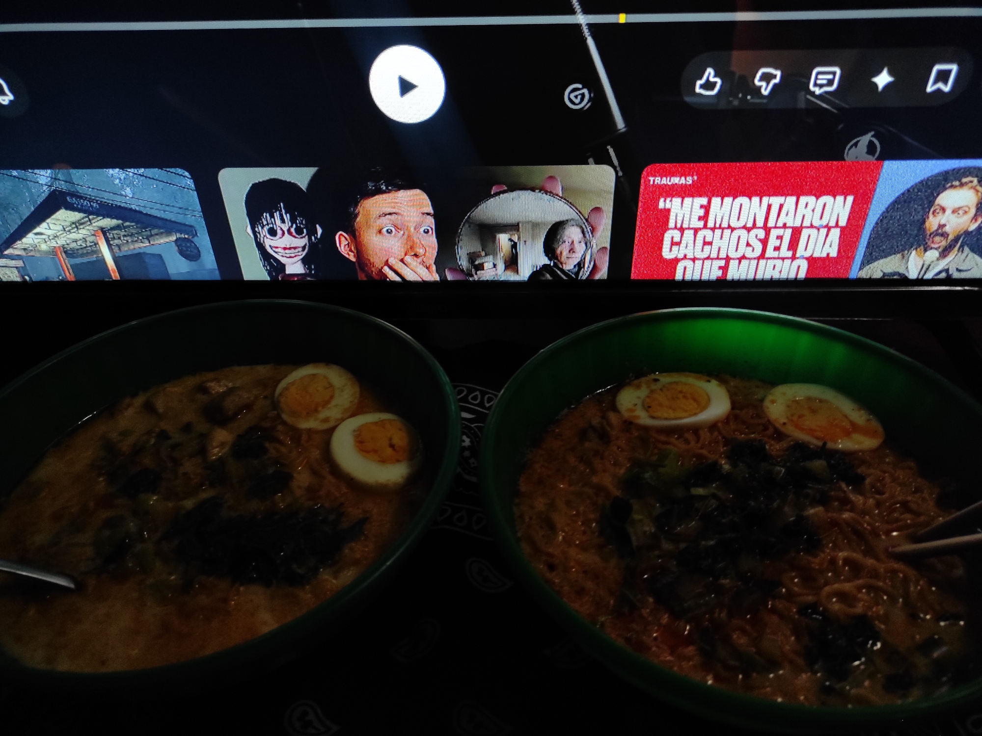 The image shows a close-up, top-down view of two bowls of ramen with hard-boiled eggs and dark green leafy vegetables. The bowls are a vibrant green. The ramen is a reddish-orange color, and the broth is visible. One bowl is positioned on the left side of the frame and the other on the right. Chopsticks are visible in the right bowl.
In the background, a television screen displays a series of video thumbnails and interface elements. The thumbnails show various images, including a distorted, creepy face with large eyes and a wide smile, a man's face looking surprised and holding a mirror, and a thumbnail with Spanish text that reads "ME MONTARON CACHOS EL DIA QUE MURIO" on a red background, next to a portrait of a man with a beard. Above the thumbnails, a play button icon is visible, along with social media interaction icons like a thumbs up, thumbs down, a comment icon, a star, and a bookmark. The overall lighting suggests the television screen is the primary light source, with the foreground bowls of ramen being dimly lit. The location is specified as Macho Muerto, Venezuela.