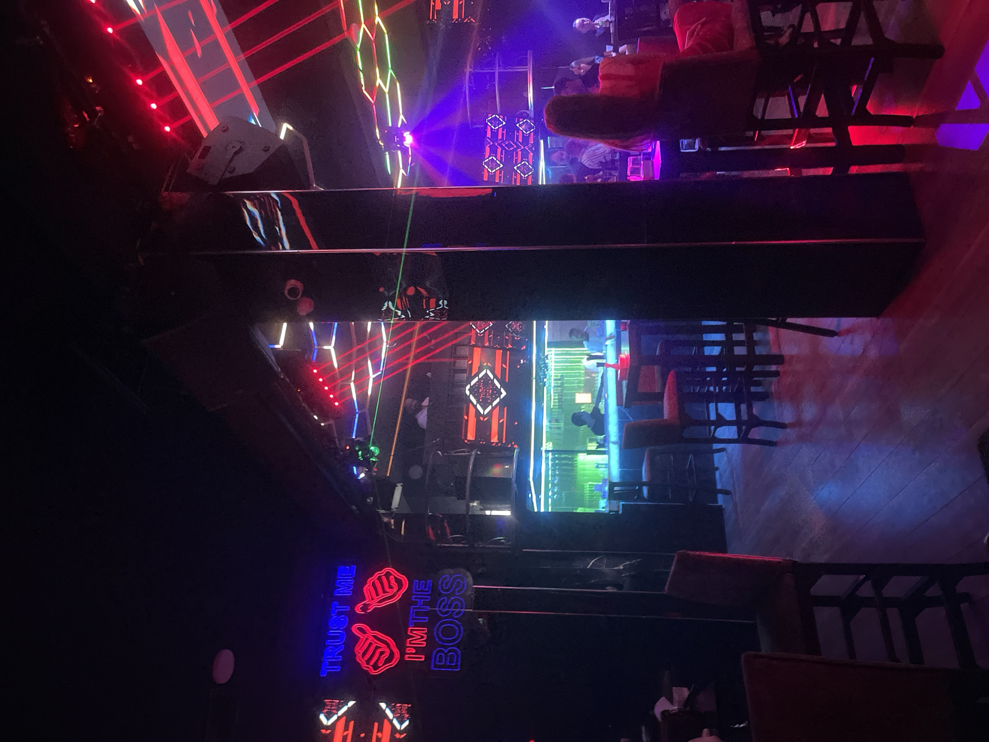 The image captures the interior of a dimly lit nightclub in Amman, Jordan. Neon signs with blue and red lights dominate the scene. A prominent blue neon sign reads "TRUST ME," accompanied by two red neon thumbs-up emojis. Below this, another blue neon sign says "I'M THE BOSS." Red and green laser lights crisscross the space, illuminating the dark environment. Several individuals are present, some seated at tables and others standing, though their faces are not clearly discernible due to the low lighting and distance. The flooring appears to be wooden planks, reflecting the colorful lights. Bar stools and tables are arranged throughout the club. A large, reflective black pillar stands in the foreground, adding to the nightclub's ambiance.