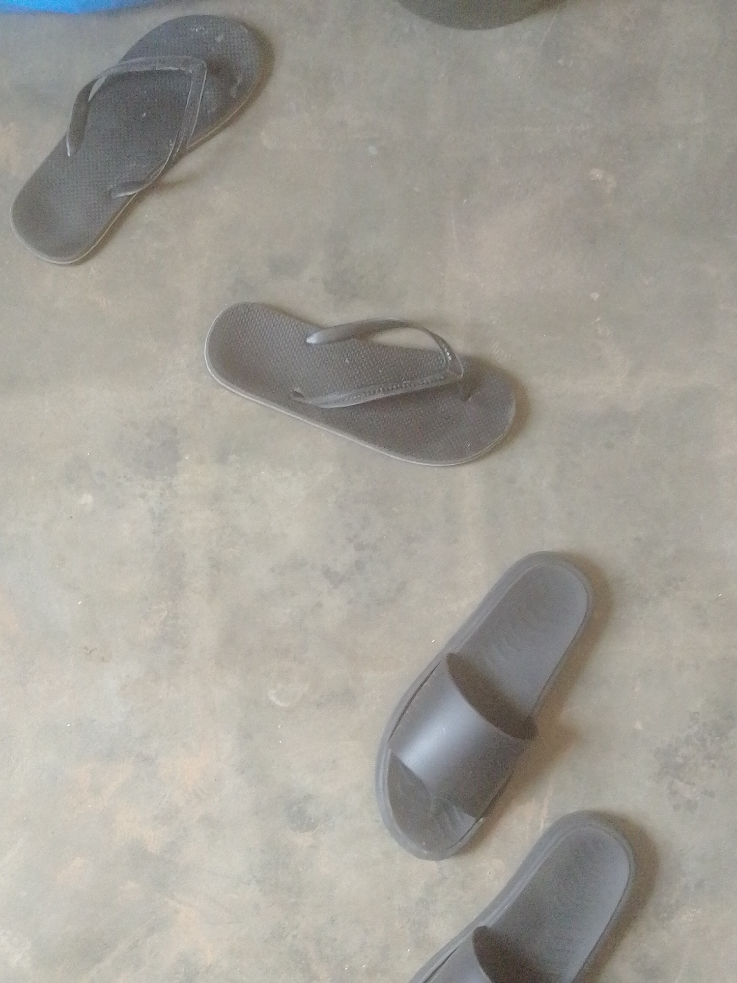 In Majemu, Nigeria, on a dull, concrete floor, four dark-colored flip-flops and slides are scattered. Two flip-flops are positioned near the top left of the image. One is open, with its strap extended upwards, and the other is closed, showing its sole. Below these, towards the center, another pair of flip-flops lies open. The lower right portion of the image features two slides, one directly above the other, both facing towards the bottom. The floor exhibits a mottled texture with variations in shade, suggesting wear and tear. There are no people or animals present in the scene.