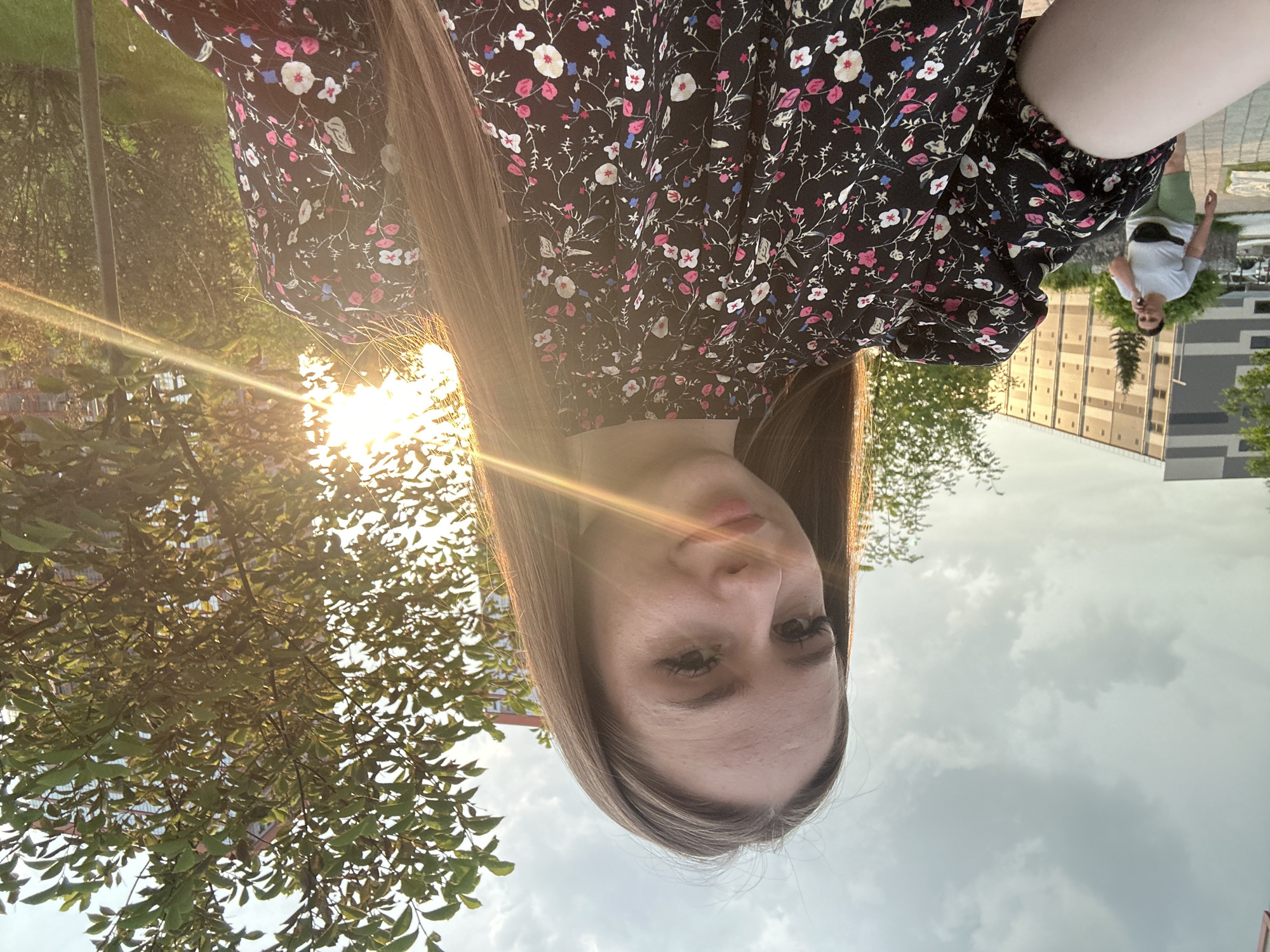 A young woman with long, brown hair is taking a selfie outdoors in what appears to be an urban environment. She is smiling and looking at the camera. She is wearing a black dress with a floral pattern featuring pink, white, and blue flowers. The background shows a modern building, trees, and a cloudy sky with the sun setting or rising, casting a warm glow and lens flares. In the background, on the left side of the image, another woman is walking and talking on a phone. The ground is paved with concrete tiles.