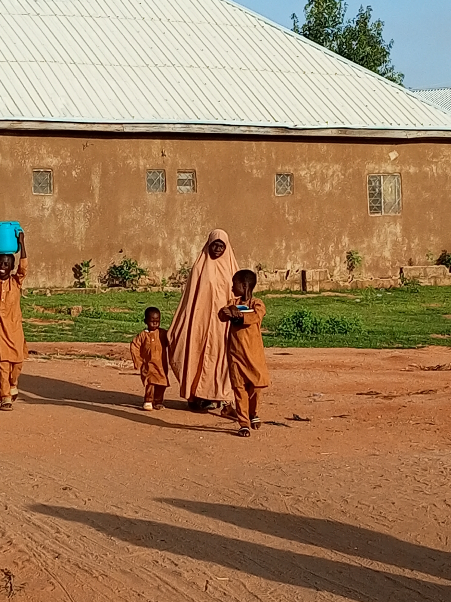 In the city of Tempure, Nigeria, a group of four people, three children and one adult woman, walk on a dirt road in front of a brown, stucco building with a white metal roof. The woman, dressed in a light brown hijab and dress, walks in the center of the group. To her right, a young boy in a brown outfit carries a blue bucket on his head. To the woman's left, a smaller child in a brown outfit walks beside her. Behind the woman and to the right, another child in a brown outfit holds a book. The building behind them has multiple small, square windows with a lattice design. The ground is a reddish-brown dirt, and patches of green grass and shrubs are visible in the background. The lighting suggests it is late afternoon, as indicated by the long shadows cast by the people on the ground.