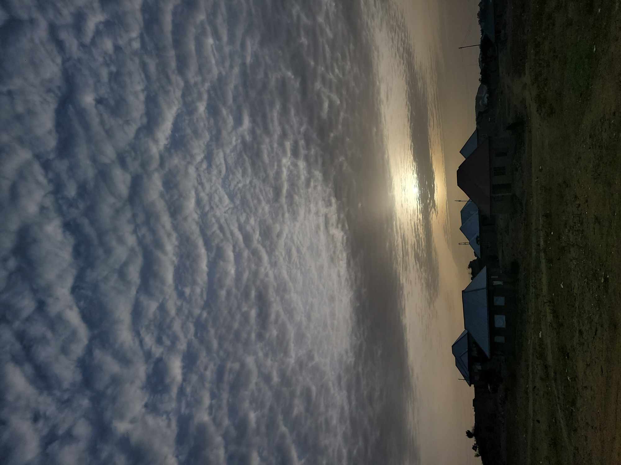 The image displays a tranquil landscape scene at either sunrise or sunset, set against a dramatic sky in Barakin Akawo, Nigeria. The sky occupies the upper two-thirds of the frame, filled with extensive altocumulus clouds exhibiting distinct wavy or ripple-like patterns, predominantly in shades of white and grey.
A bright, intense sun is visible near the horizon, partially obscured by a lighter band of clouds, creating a strong glare and casting a warm, golden-yellow glow across the central portion of the sky. This backlighting heavily silhouettes the elements below.
Along the horizon, a row of several houses is visible, their dark forms sharply contrasted against the illuminated sky. These appear to be single-story structures with sloped roofs. In the foreground and to the side of the houses, there is a patch of green and brownish-green land, likely a grassy field or undeveloped terrain. Faint details of utility poles or wires can be discerned above the distant houses.
No people or specific activities are present in the image, highlighting the quiet, natural beauty of the moment. The low-angle sunlight and the intricate cloud formations create a serene and contemplative atmosphere, characteristic of the early morning or late evening. No visible text is present.