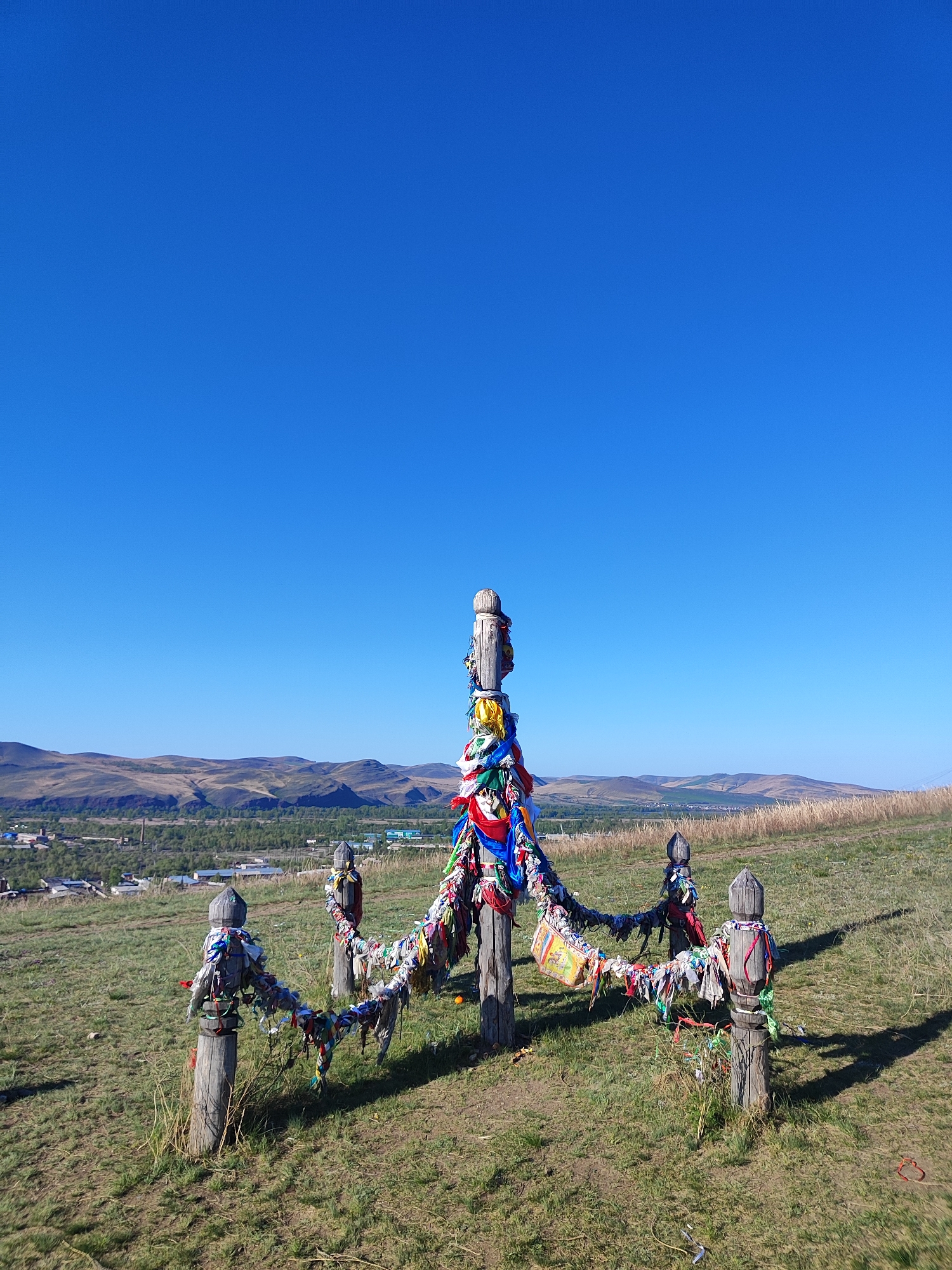 The image shows a structure made of carved wooden posts standing in a grassy field. The posts are connected by ropes or strings that are adorned with colorful ribbons and pieces of fabric. In the background, there is a town or village surrounded by rolling hills under a clear blue sky. The image was taken during the day.