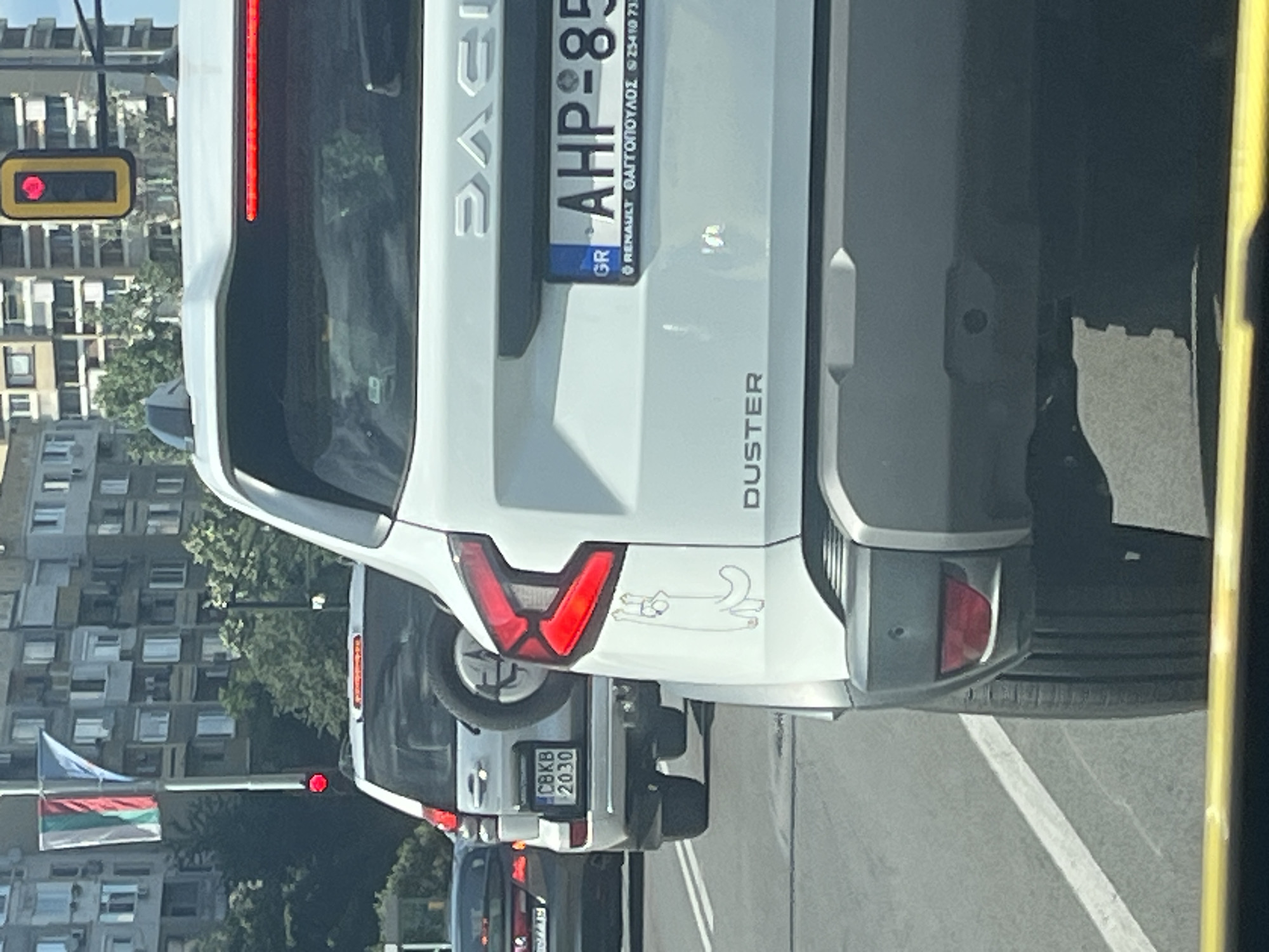 In Sofia, Bulgaria, on a clear day, cars are stopped at a traffic light. A white Dacia Duster is in the foreground, with a sticker of a white cat with its arms up on its rear passenger side door. The license plate reads "AHP-85" with "GR" on the left, indicating a Greek registration. To the left of the Duster, a silver SUV is stopped, with a spare tire mounted on its rear. Its license plate reads "CBKB 2030". In the background, apartment buildings line the street, and a traffic light with a red signal is visible. The image captures a moment of urban traffic in Sofia.
