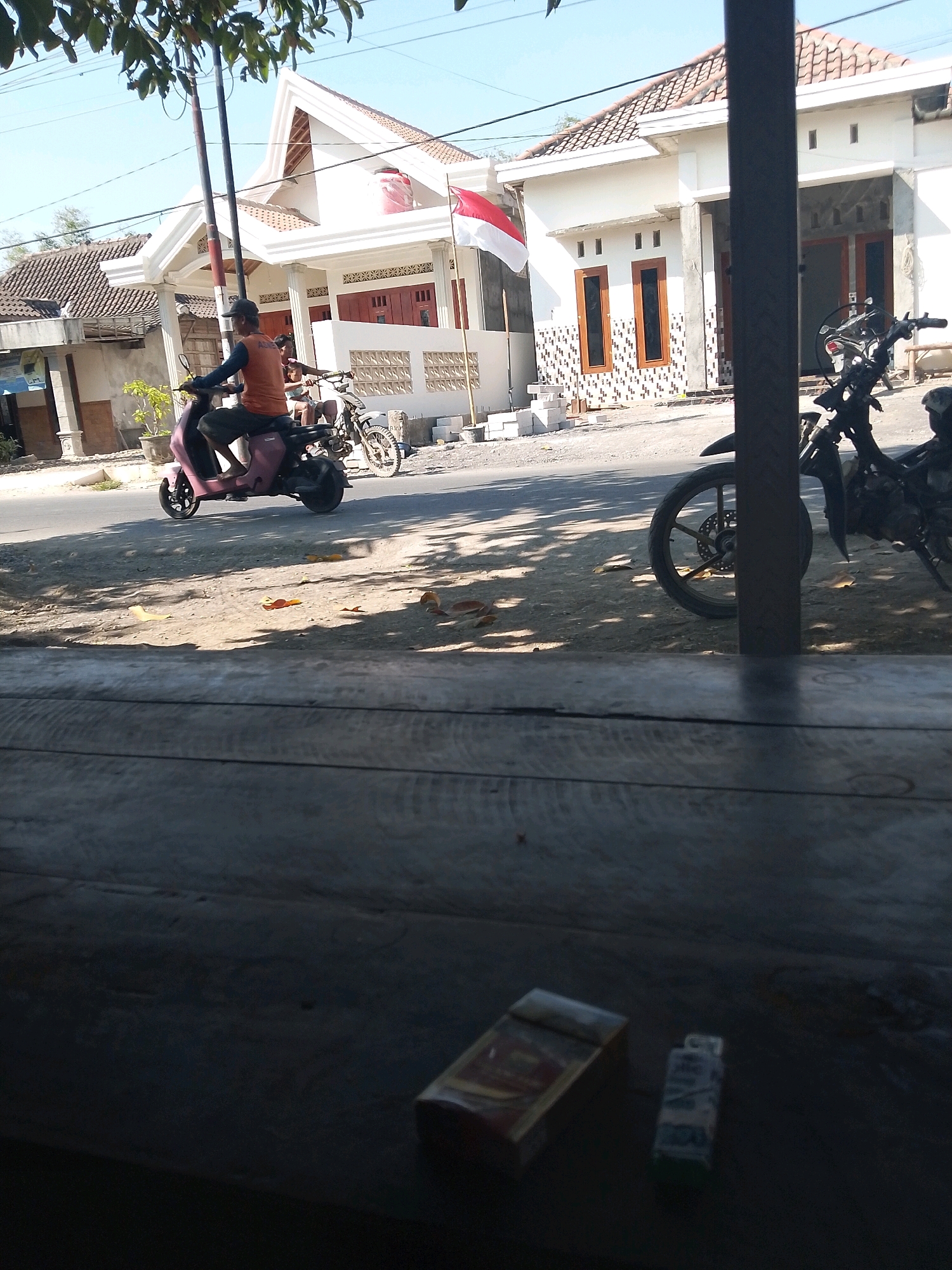 An outdoor, eye-level medium shot shows a person riding a pink scooter in what appears to be the city of Wadang, Indonesia. The scooter is moving on a paved road, and a second motorcycle is parked on the side of the road to the right. A wooden surface occupies the foreground of the image, and a pack of cigarettes and a lighter are visible on the wooden surface. The background includes residential buildings with red-tiled roofs and white walls, along with a flagpole with an Indonesian flag. The sky is clear and blue.