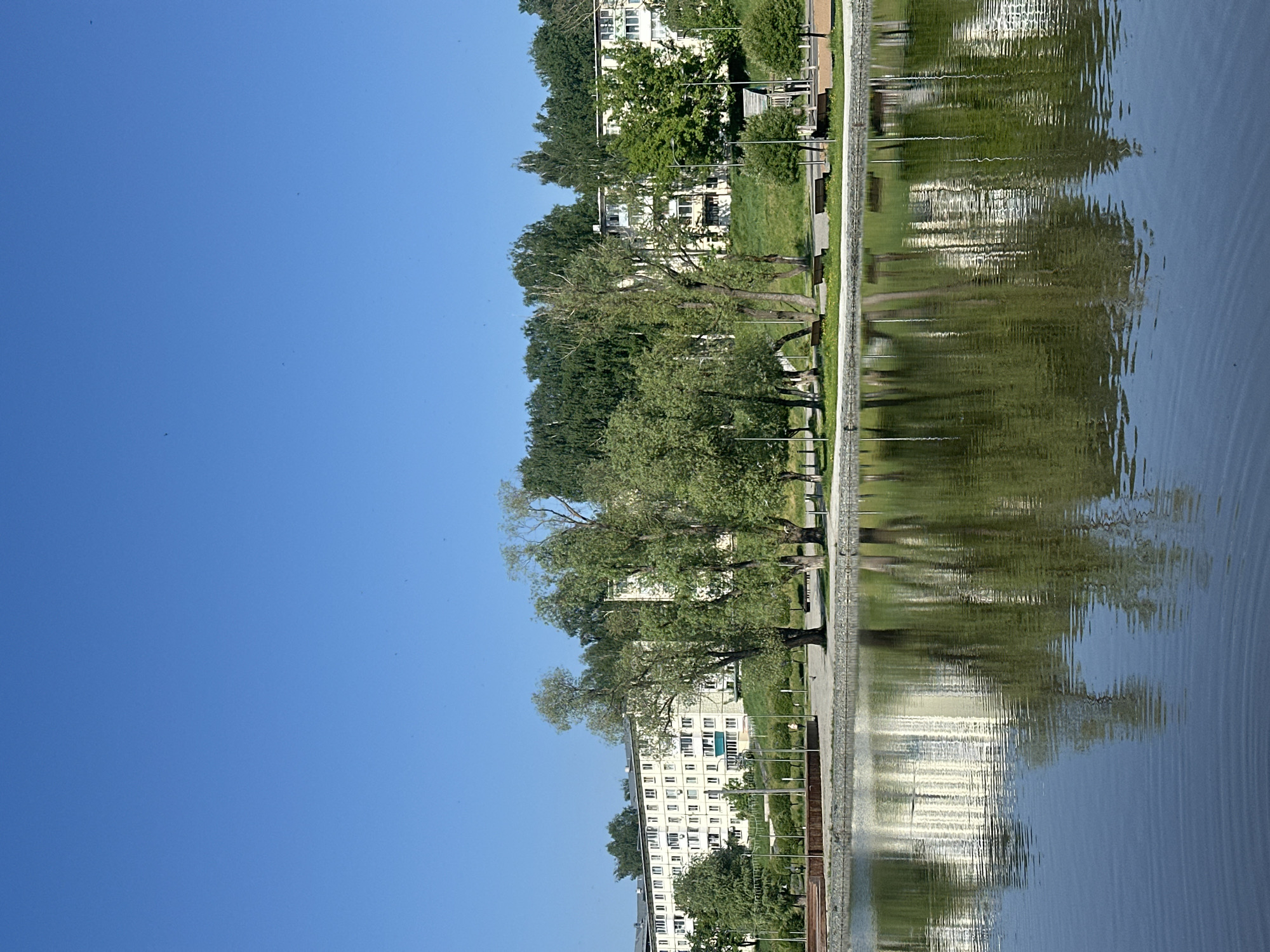 The image depicts a park scene in Votkinsk, Russia, as reflected in a body of water. The sky is a clear blue, occupying a significant portion of the image. The reflection shows trees, a building, and park infrastructure along the water's edge. The water's surface shows ripples, distorting the reflected image. A walkway borders the water. Benches and lampposts are situated along the path. The building has multiple stories and rows of windows. Green grass and trees surround the building.
