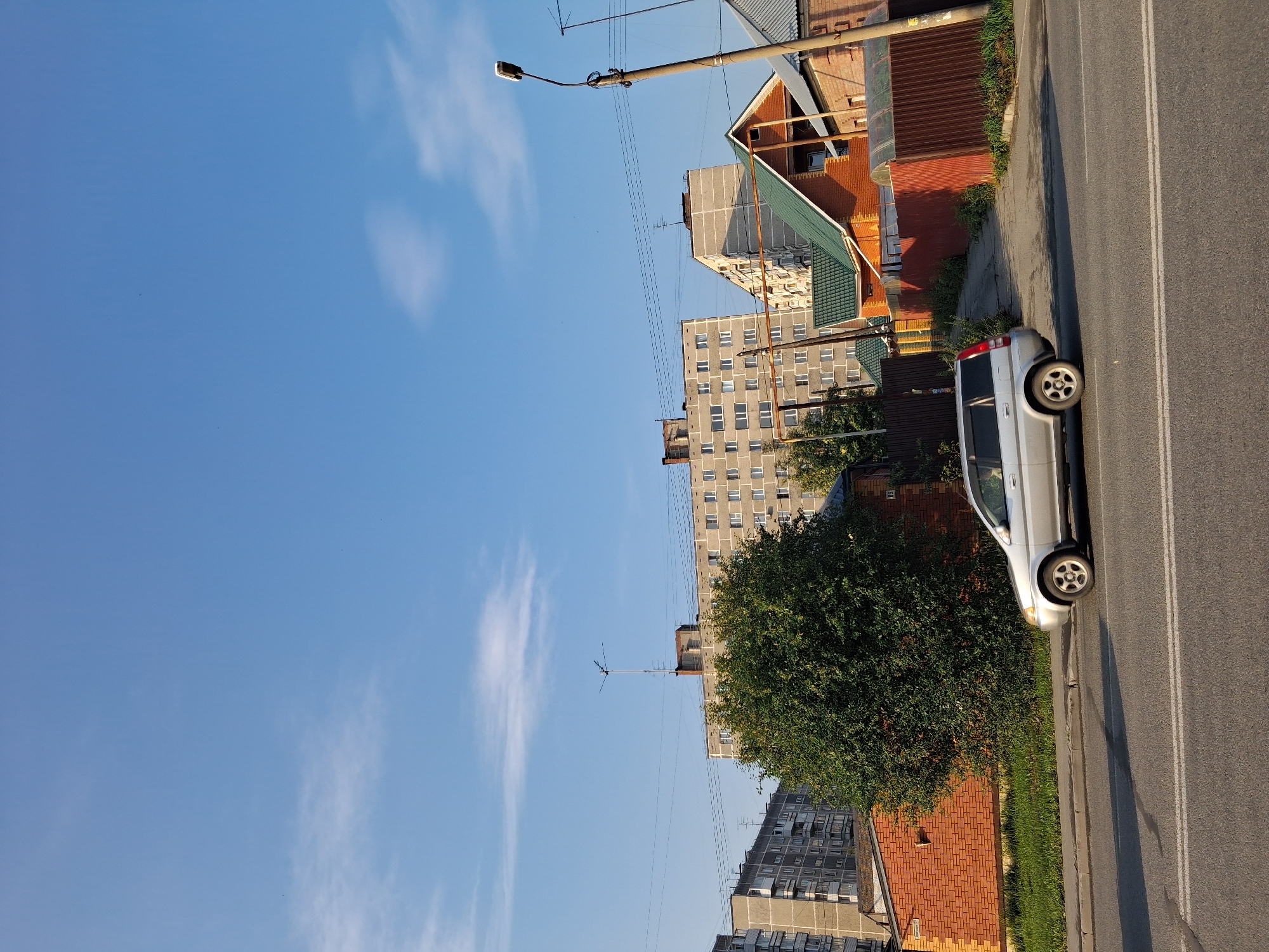 The image shows a silver Honda CR-V driving on a paved road in front of a brick wall and a large green tree. In the background, there are multi-story apartment buildings and smaller residential houses with green roofs. A lamppost is visible on the right side of the image. The sky is clear and blue with a few wispy clouds. The scene appears to be a typical urban environment in Novosibirsk, Russia, likely during daytime given the sunlight and shadows.